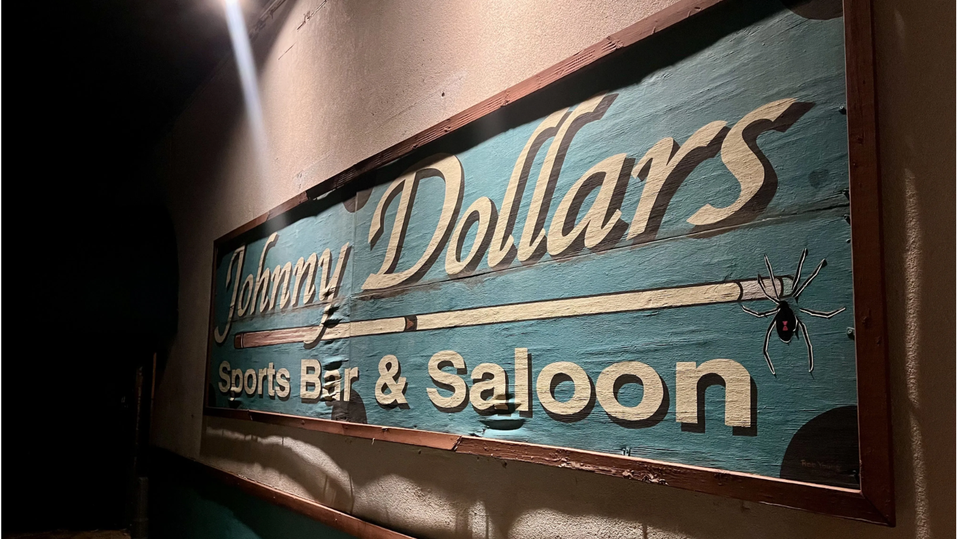 Night view of a weathered wooden sign for Johnny Dollars Sports Bar & Saloon with a blue background and a black widow spider illustration on the right edge.
