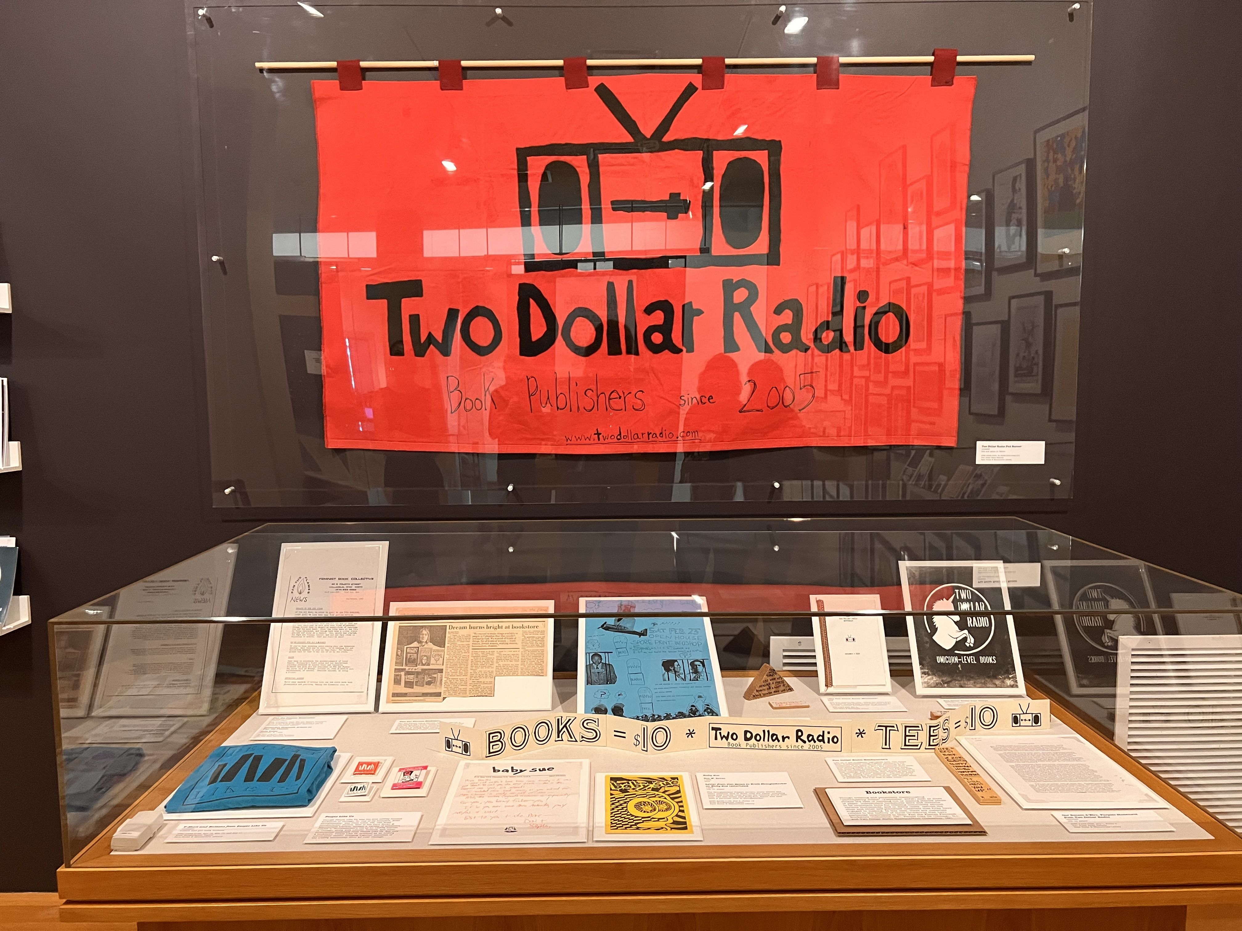 Red banner in a glass display case features a black, cartoon TV icon with antenna. Large text reads \"Two Dollar Radio\" and a smaller line says \"Book Publishers since 2005\" underneath.