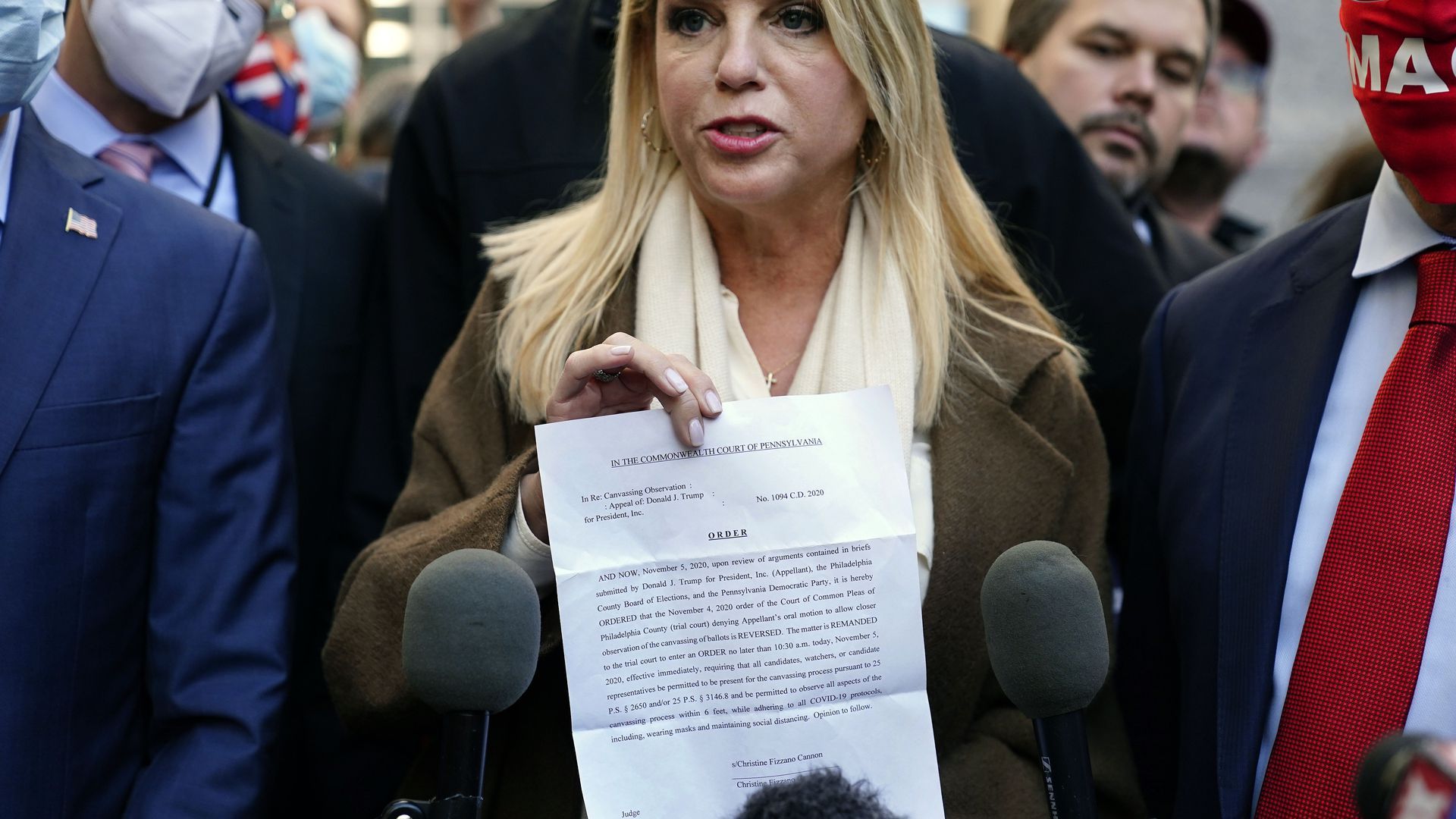 A blonde woman holds a sheet of paper in front of two microphones, with a crowd standing around her mostly of men wearing suits