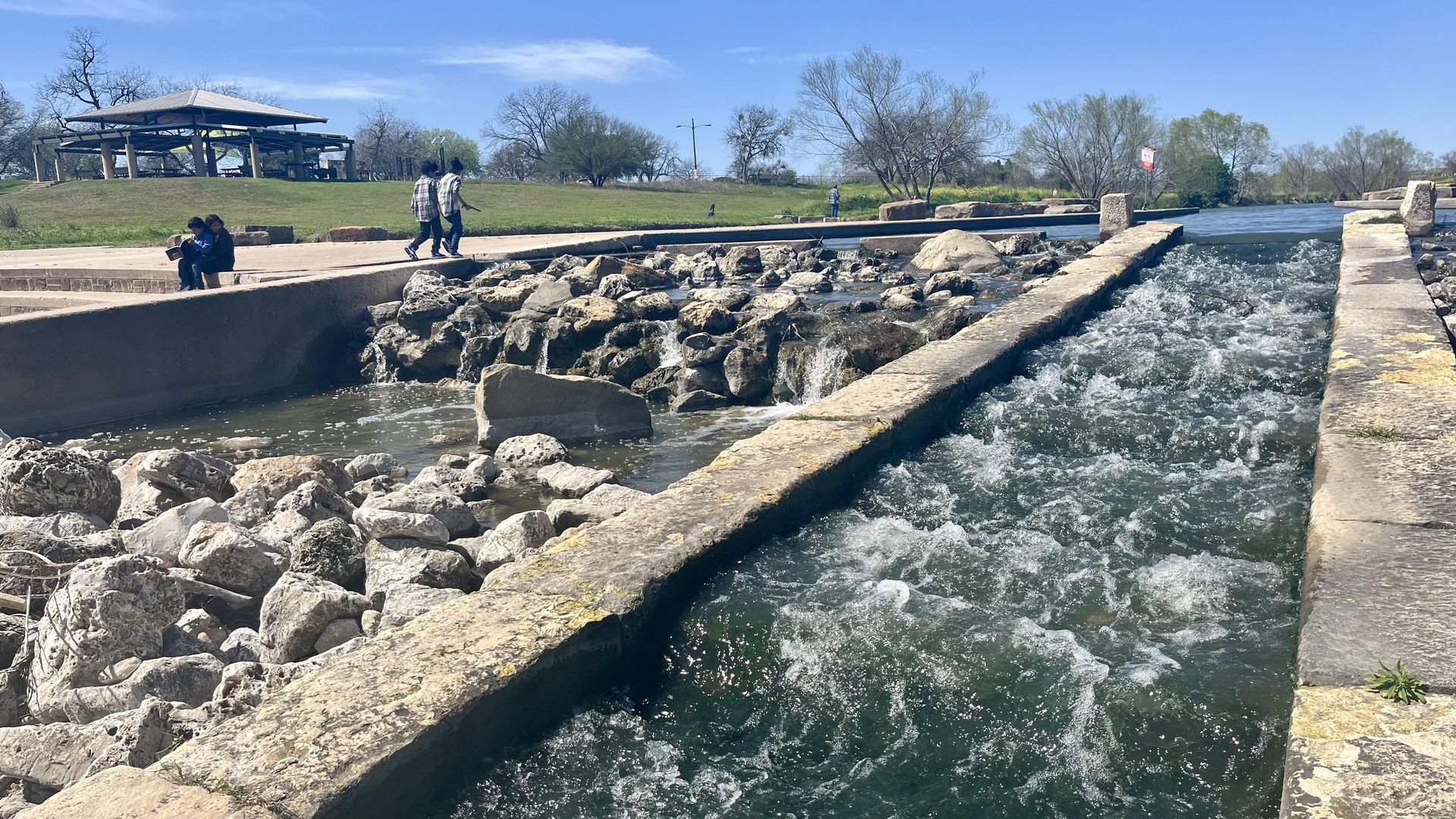 The San Antonio River water rushes through a dam with a park pavilion and people walking by in the background.