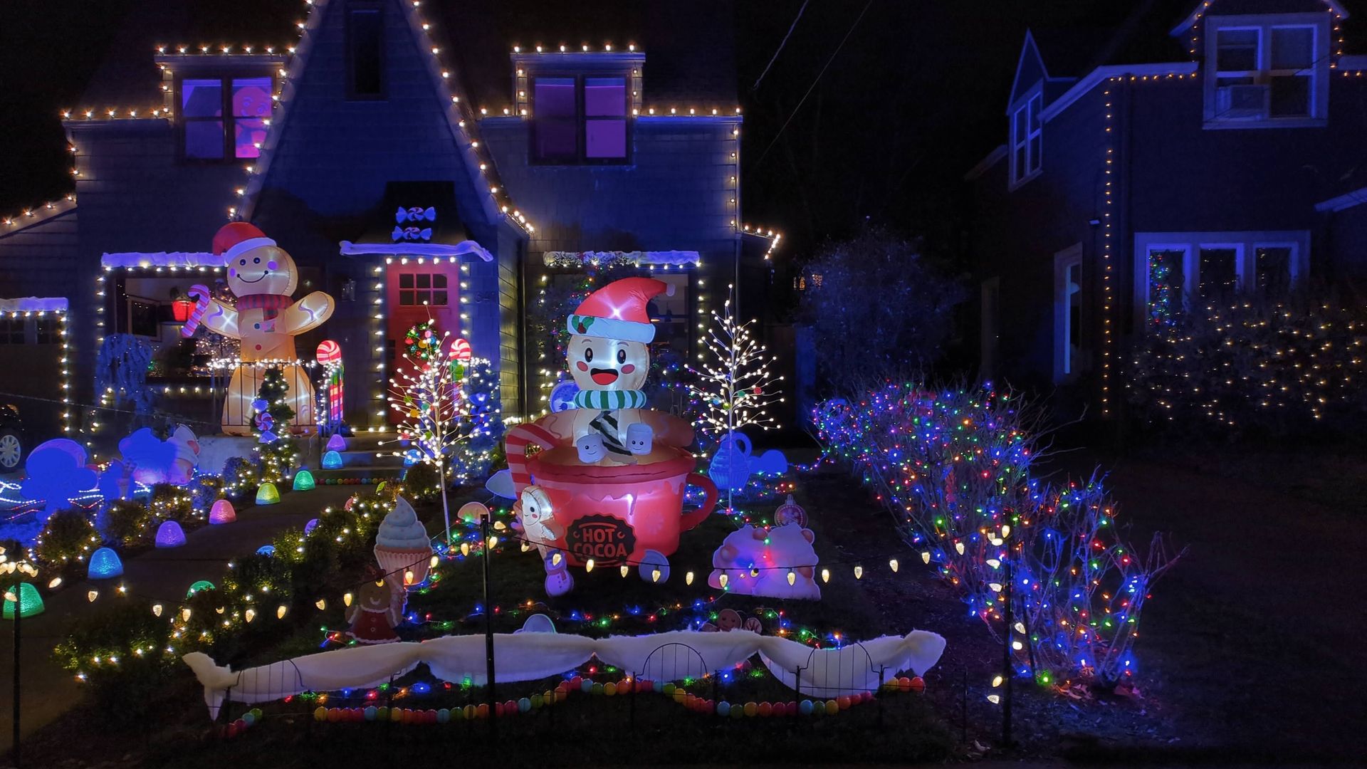 House decorated with white, blue, and multicolored Christmas lights, inflatable gingerbread man, snowman in hot cocoa cup, candy cane decorations, and small lighted trees at night.