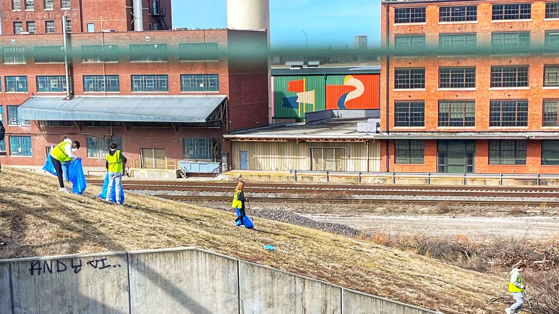 Several workers in yellow safety vests pick up litter on a grassy embankment beside a railway, carrying blue trash bags, with brick industrial buildings and a bright mural in the background.