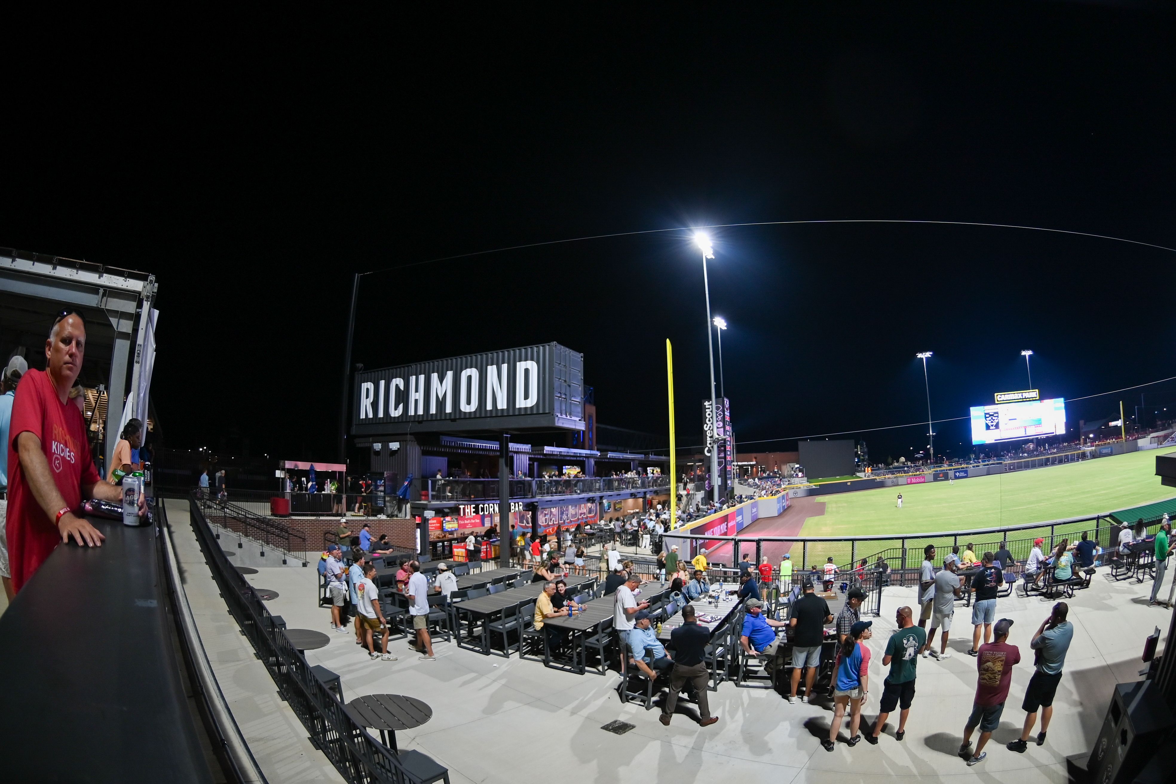 Nighttime baseball stadium in Richmond with a crowd and a man in a red shirt at the left railing, a large "RICHMOND" sign, bright floodlights, and a curved light streak across the sky.