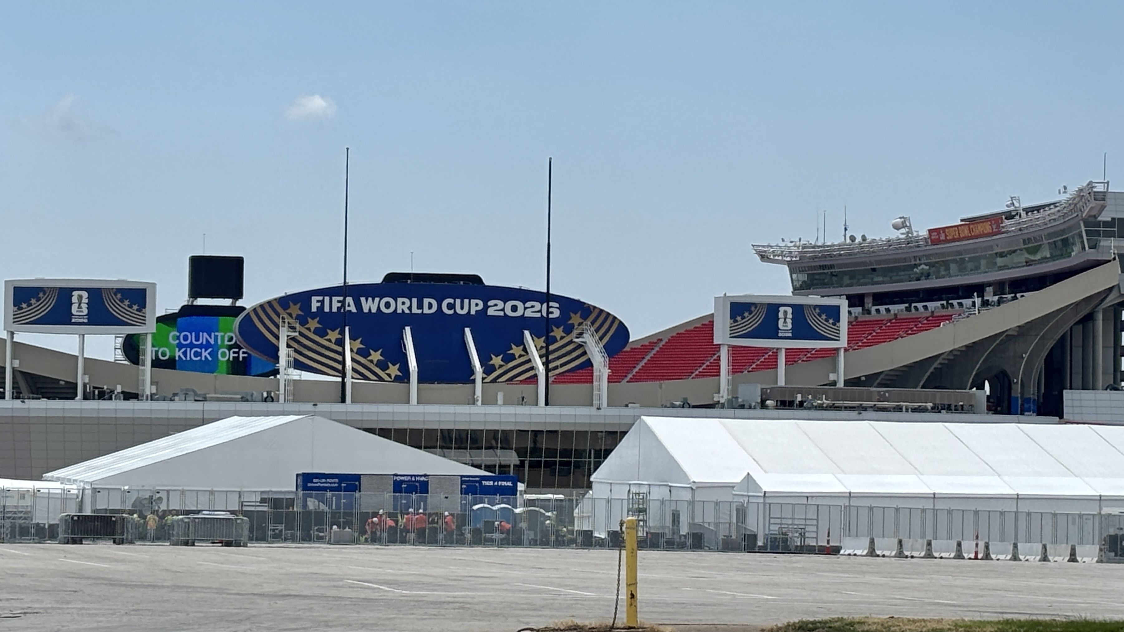 Outdoor stadium with a large blue sign reading FIFA WORLD CUP 2026 and gold stars, red seating on the right, white barriers, and white tents in the foreground under a clear blue sky.