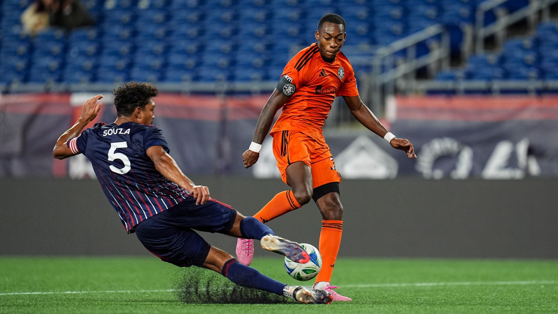 Two soccer players vie for a ball, one in an orange jersey and another in blue.