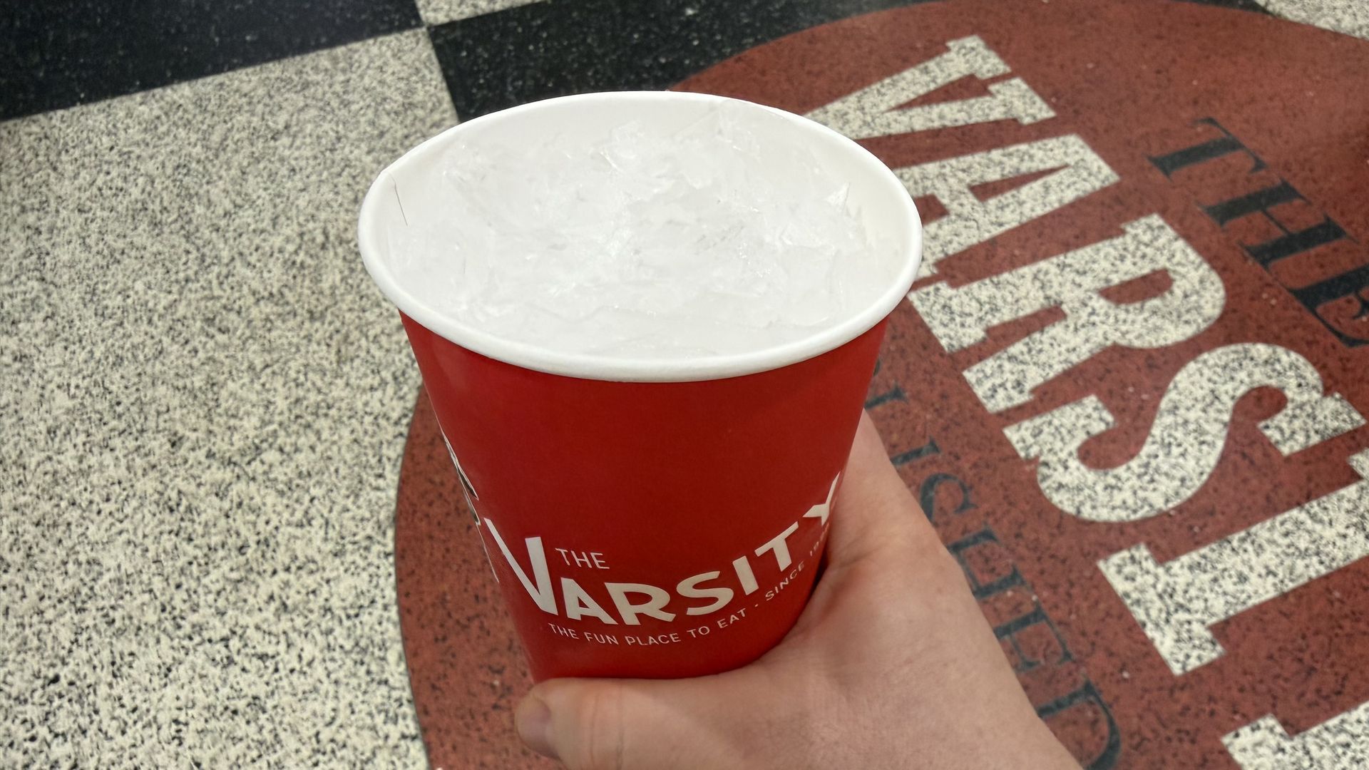 A hand grips a red disposable cup filled with ice, labeled "The Varsity." The cup sits against a speckled gray ground with bold white letters on a red surface in the background.