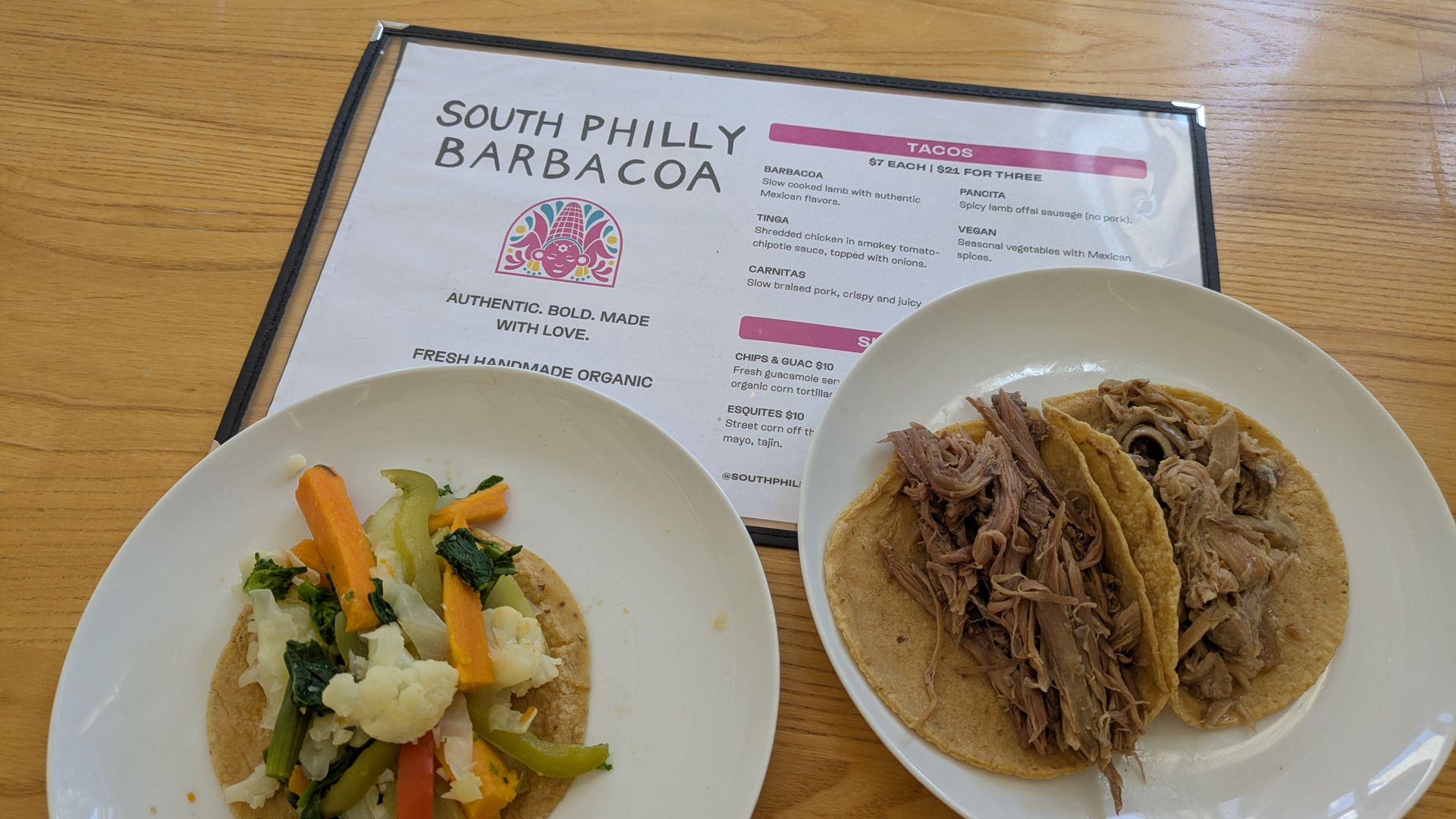 Two tacos on white plates atop a wooden table; left topped with sautéed vegetables, right with shredded barbacoa. A menu in the background reads "SOUTH PHILLY BARBACOA".