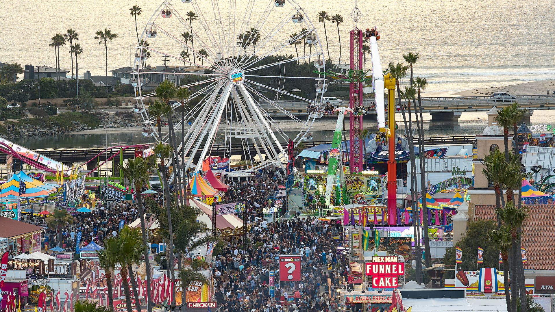 Crowded fairgrounds by the beach with a large white Ferris wheel, palm trees, colorful tents, and signs for funnel cake; the ocean glistens in the background.