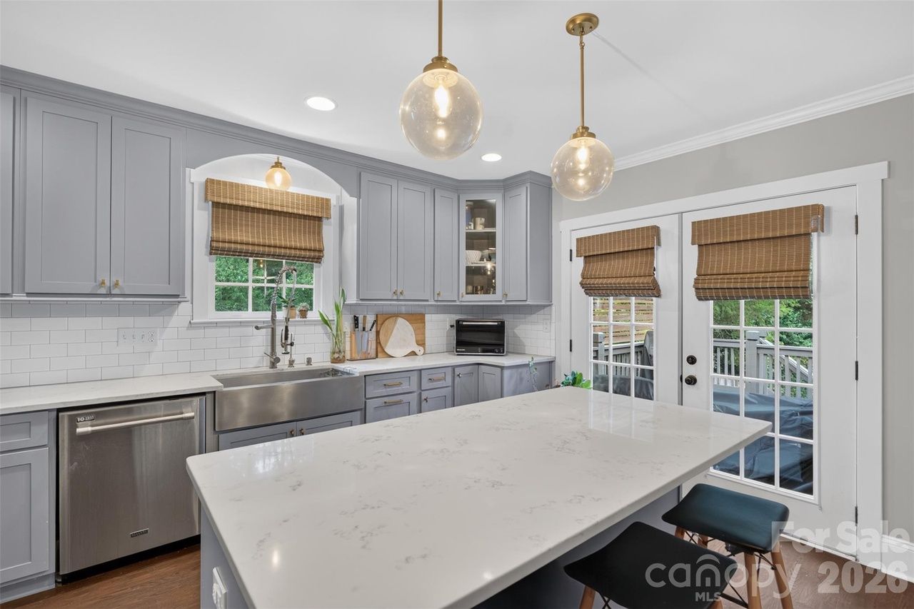 Modern gray kitchen with a white marble island, stainless steel appliances, and white subway tile. Gray cabinetry, two glass globe pendant lights, bamboo blinds, and French doors opening to a deck.