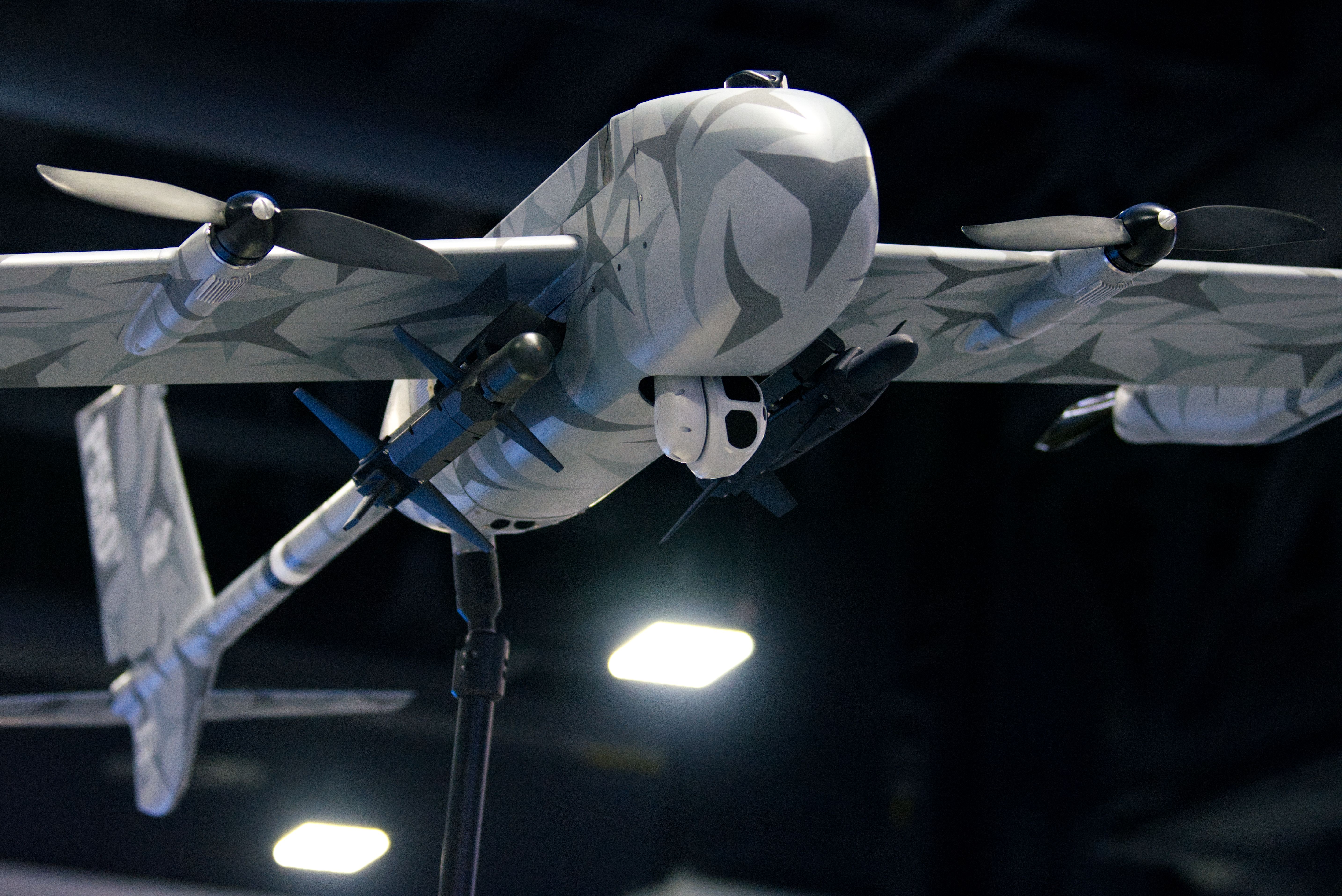 A grey-camouflaged drone is seen about a show floor booth. Propellors and munitions are seen on it,