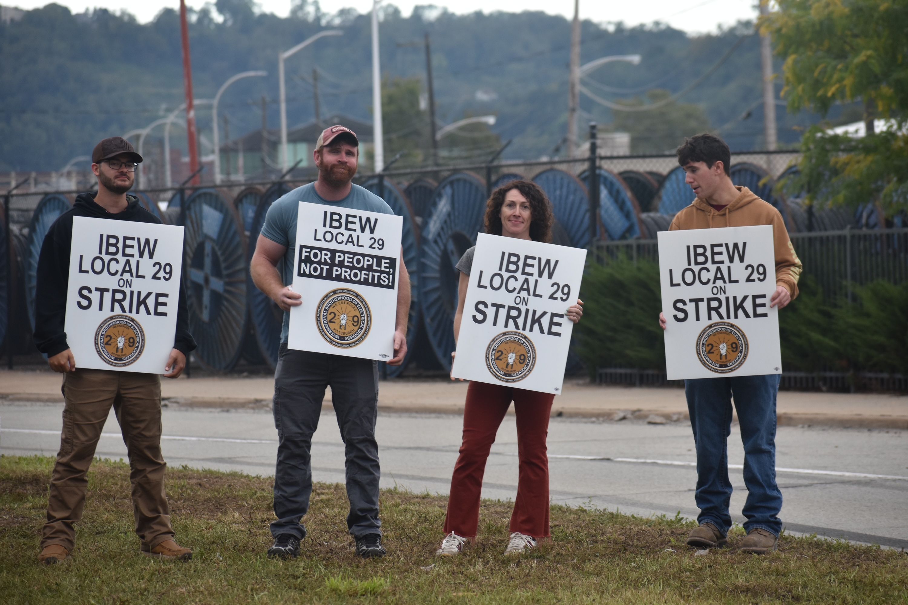 Four protesters hold white signs reading "IBEW LOCAL 29 ON STRIKE" on a sidewalk; left to right: man in a black hooded sweatshirt, man in a blue shirt, a woman with curly hair and a man in a tan hooded sweatshirt, with an industrial fence behind.