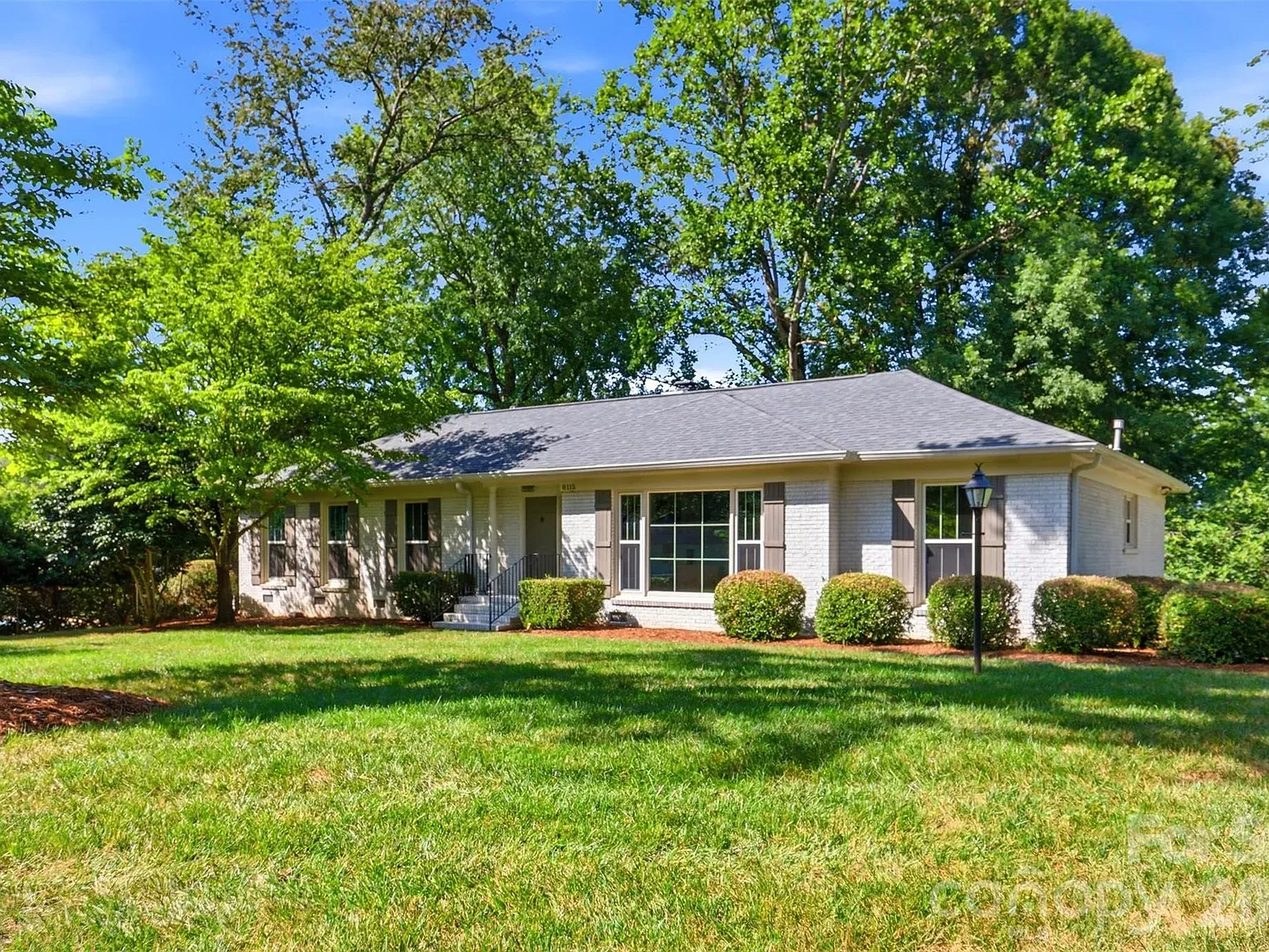 Single-story white brick house with a gray roof, front steps and porch, large front window with shutters, trimmed hedges, green lawn, and tall trees against a blue sky.