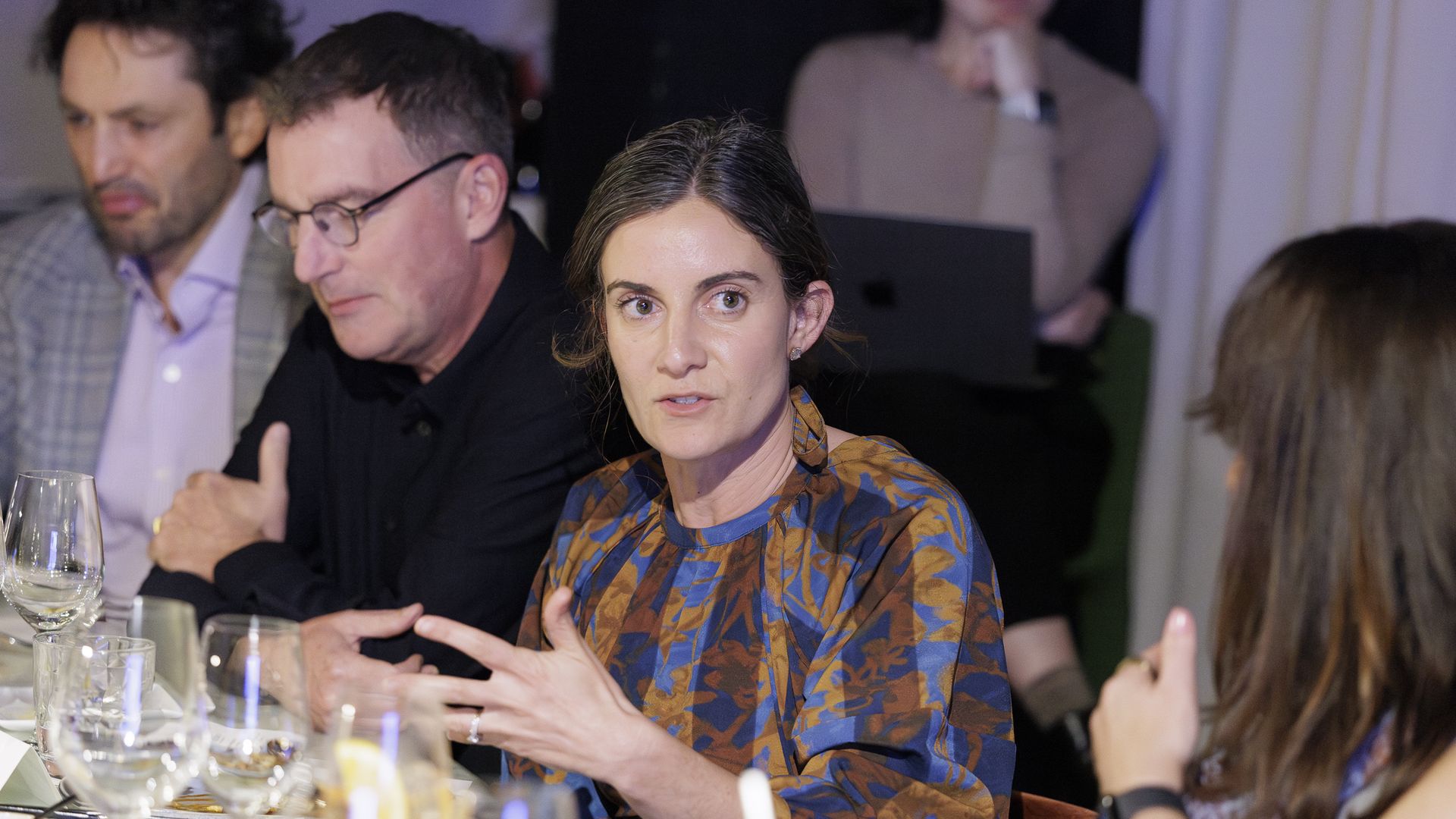 A woman in a blue-brown patterned blouse speaks and gestures at a dinner table; two men listen nearby, with wine glasses, plates, and a laptop visible in a softly lit event.