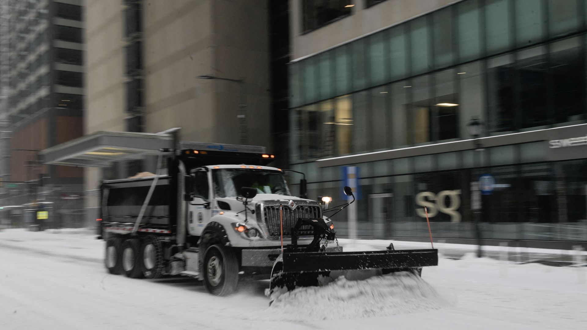 A snowplow clears snow on a roadway in front of a several high-rise buildings. 
