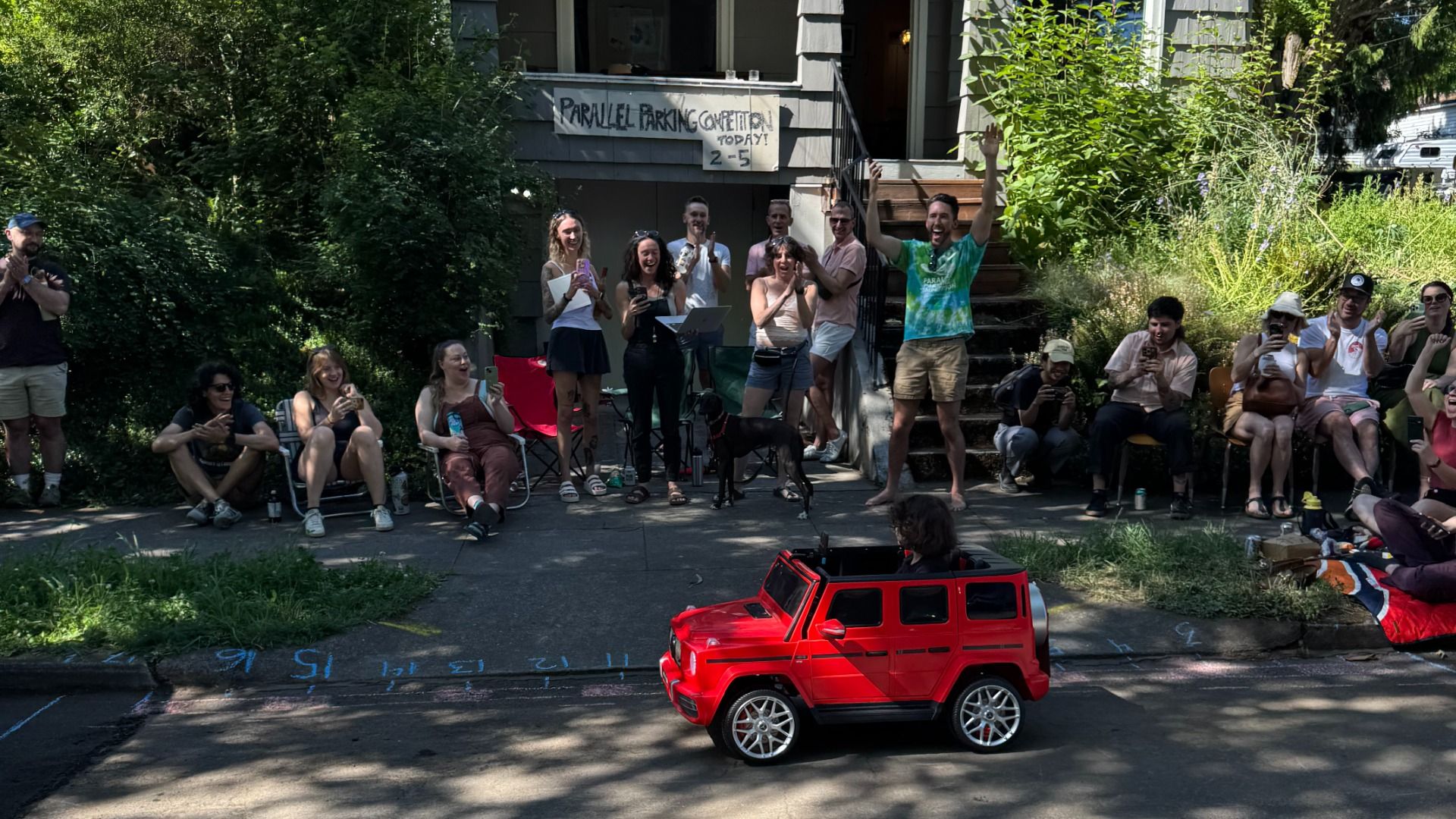 A crowd gathers outside a Portland home for a parallel parking competition as a child drives a small red toy SUV along the street.