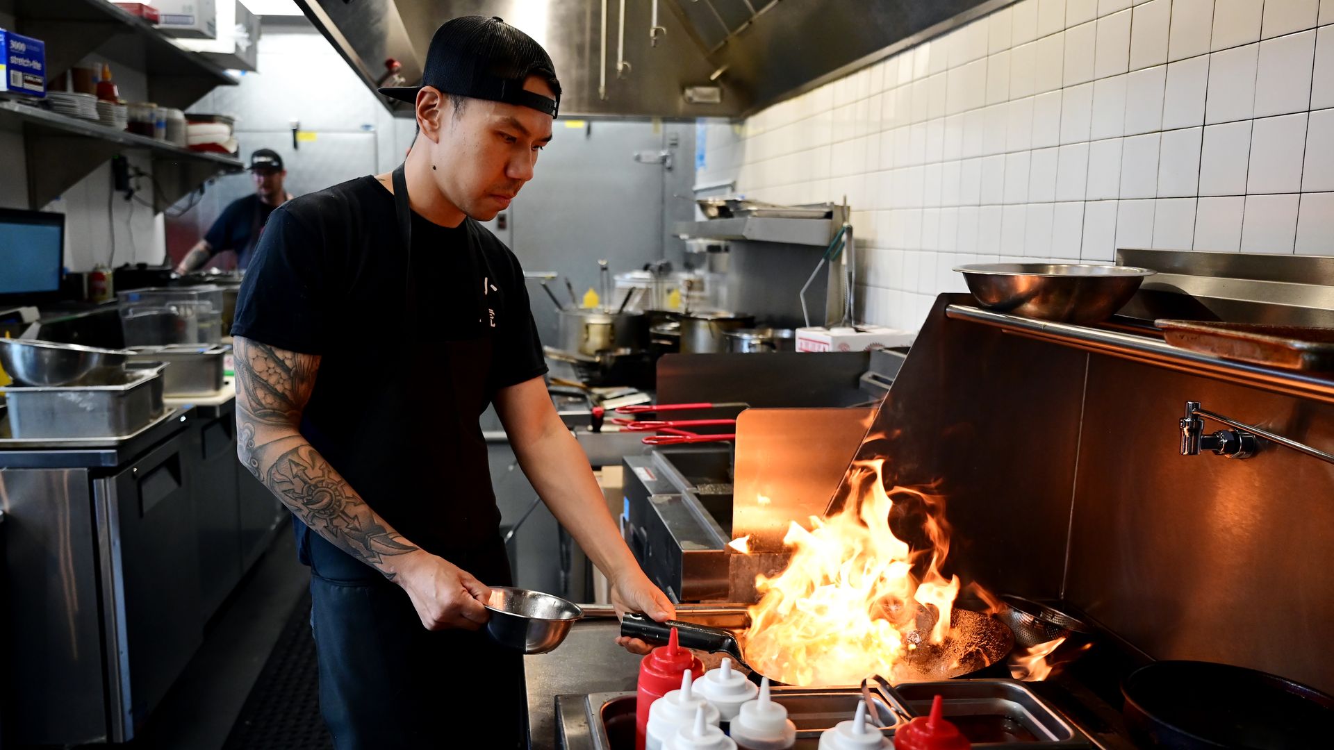 Chef in a black cap and shirt grills over open flames in a busy commercial kitchen, holding a pan in one hand and tongs in the other as stainless steel equipment and white tiled walls loom behind.