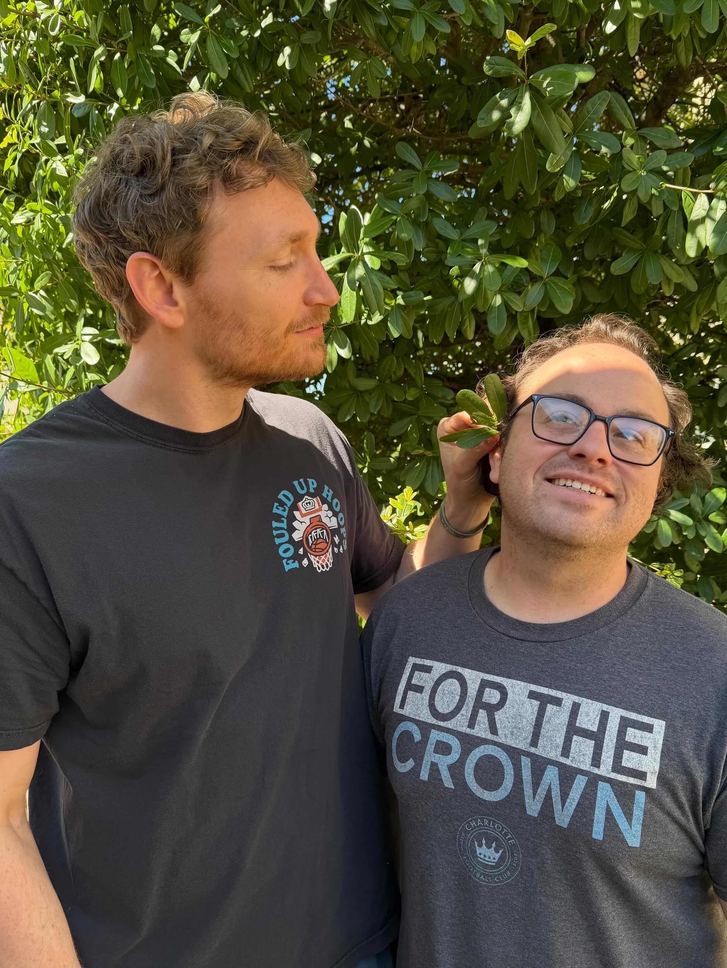 Two men posing outdoors in front of dense green leaves. Left man with short light brown hair and stubble looks at the smiling man on the right, who wears glasses and a gray shirt reading FOR THE CROWN.