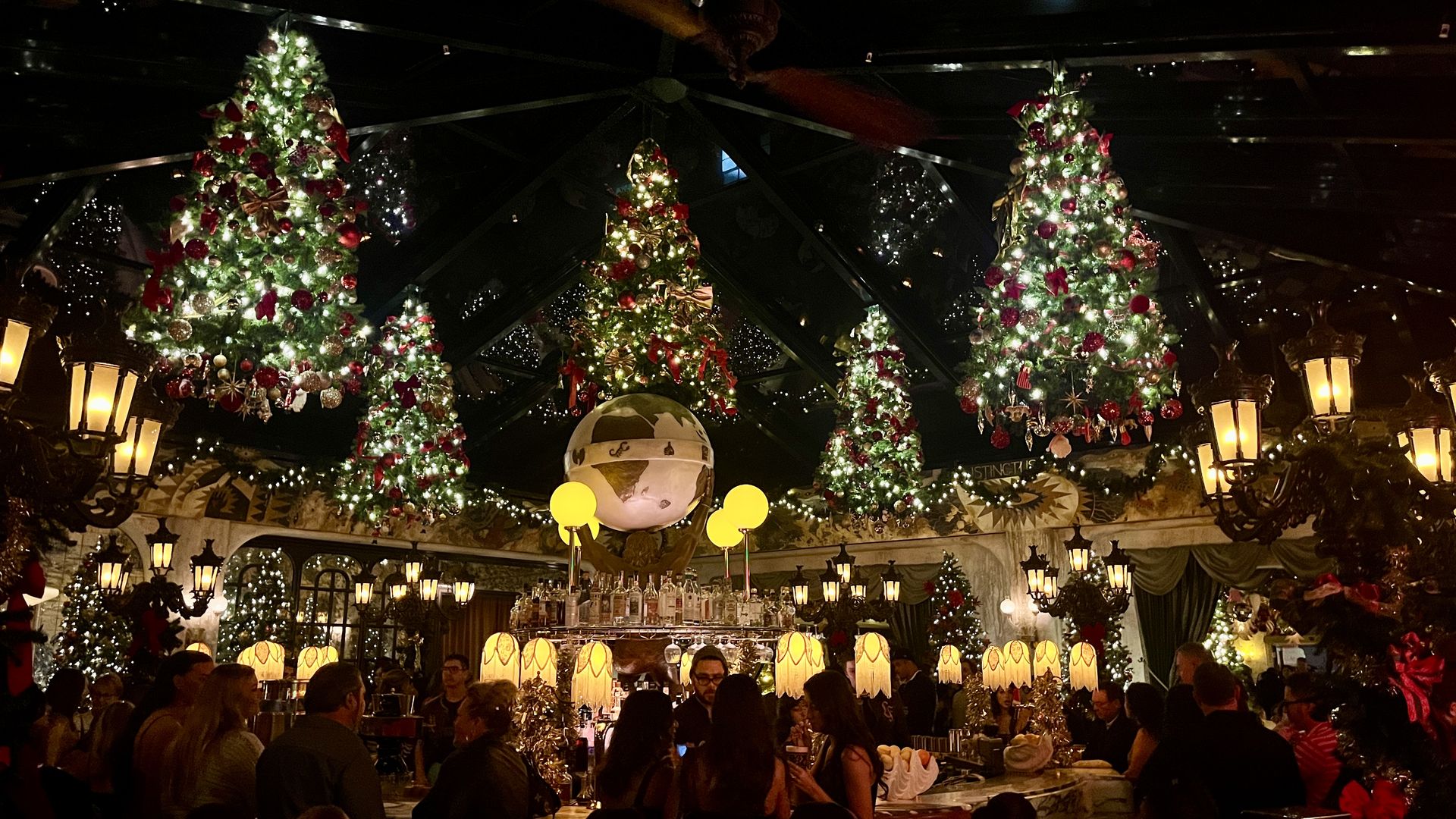 Dimly lit festive bar with five upside-down Christmas trees hanging from ceiling, decorated with red and gold ornaments, crowd of people, warm glowing lamps, and a globe centerpiece.