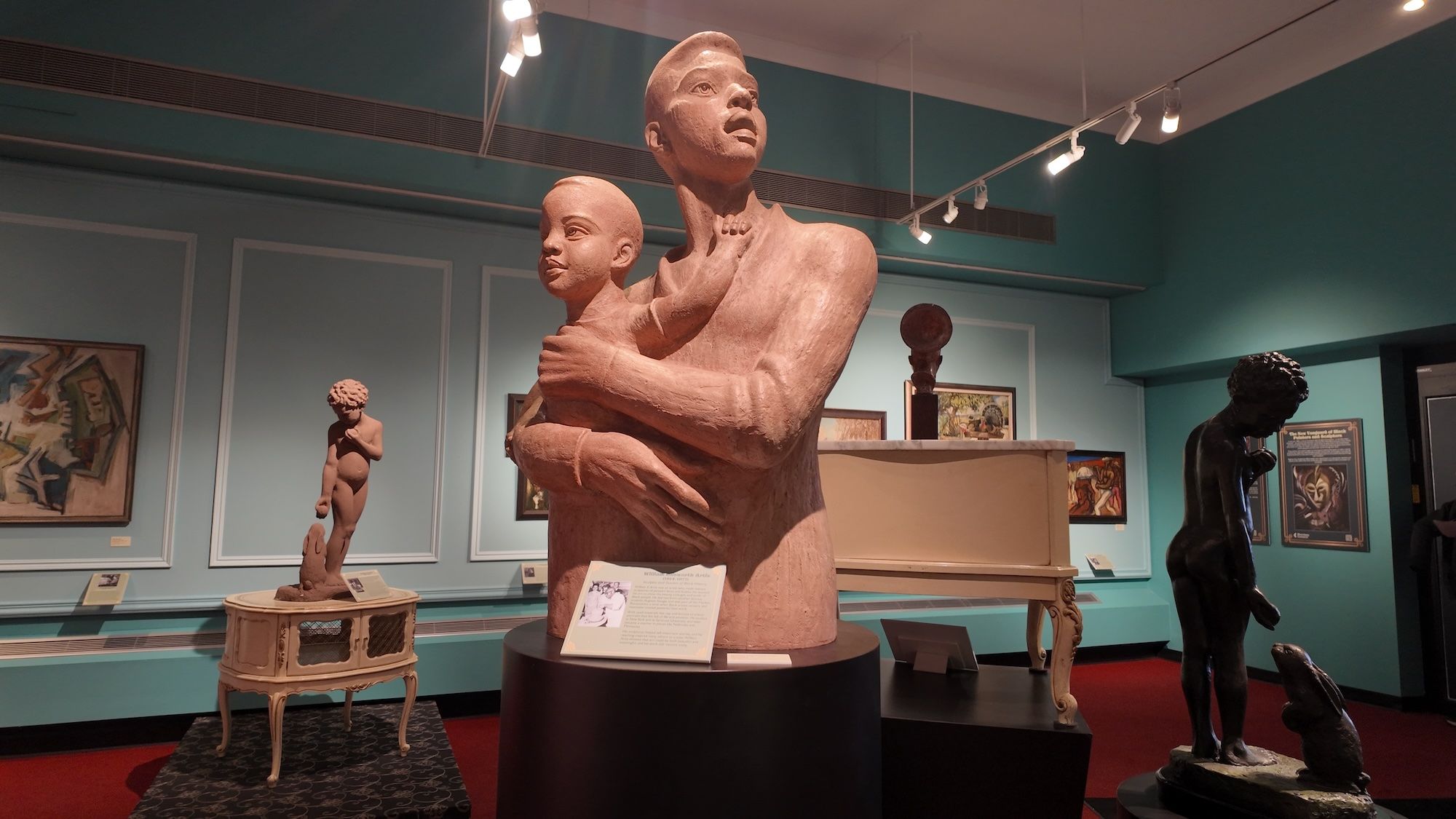 Museum room with several sculptures including a large terracotta-like statue of a person holding a child in the center, a smaller nude child statue on a table to the left, and a dark statue of a child with a rabbit on the right.