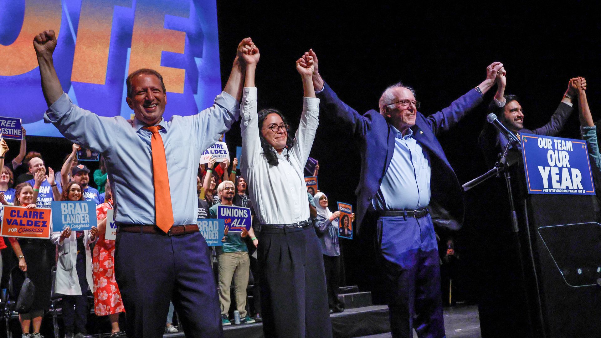 Diverse political candidates on a campaign stage raise their hands in victory; a large blue and orange banner with "VOTE" behind them, while a cheering crowd holds signs like "Our Team Our Year".