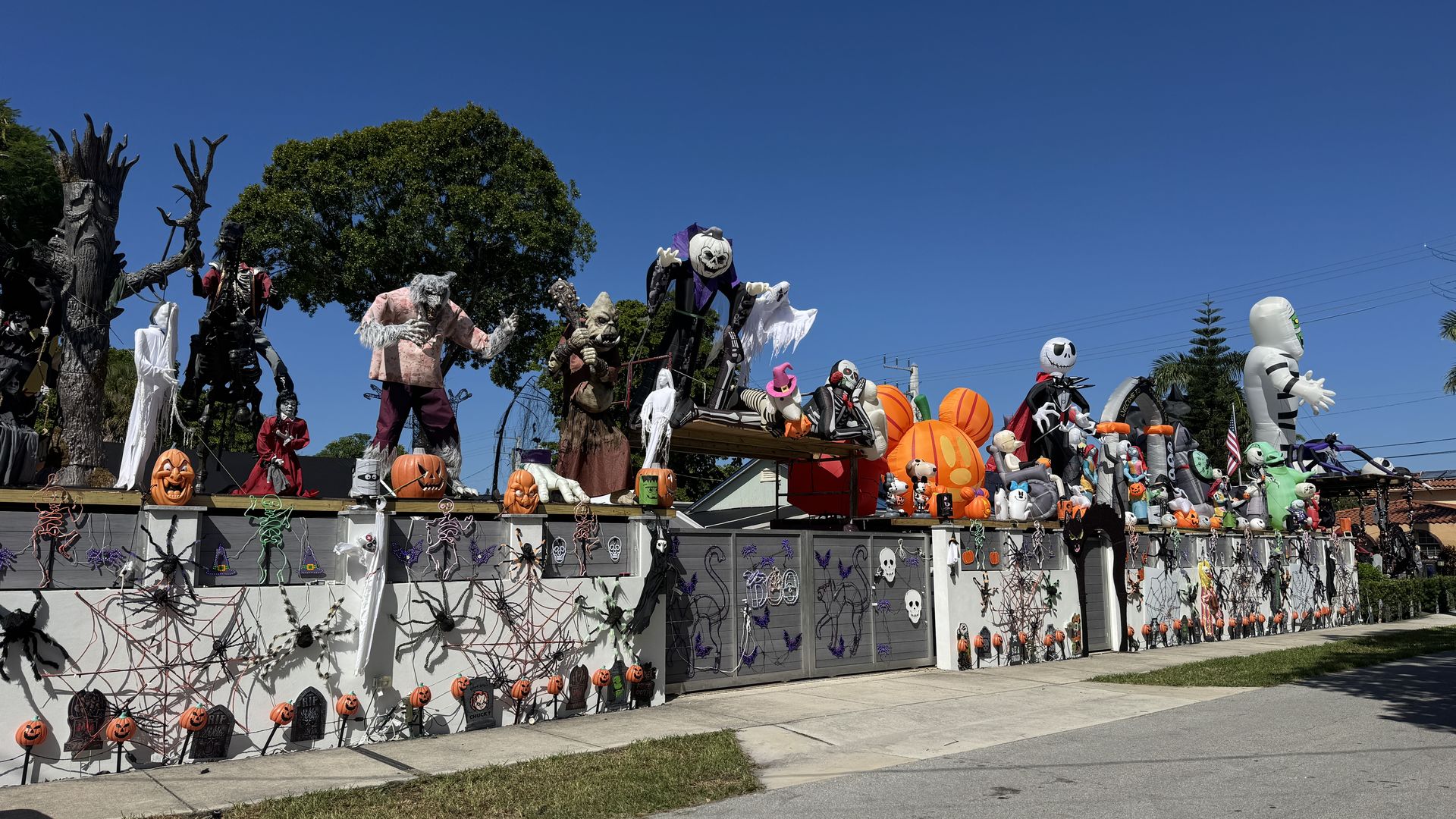 A line of Halloween decorations atop a wall, including carved pumpkins, inflatable ghosts and skeletons, and various spooky figures under a clear blue sky.