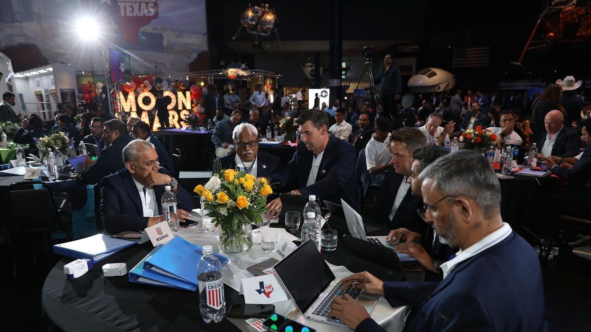 Men in nice suits sit around a table, looking thoughtful. Some have computers, some have papers.