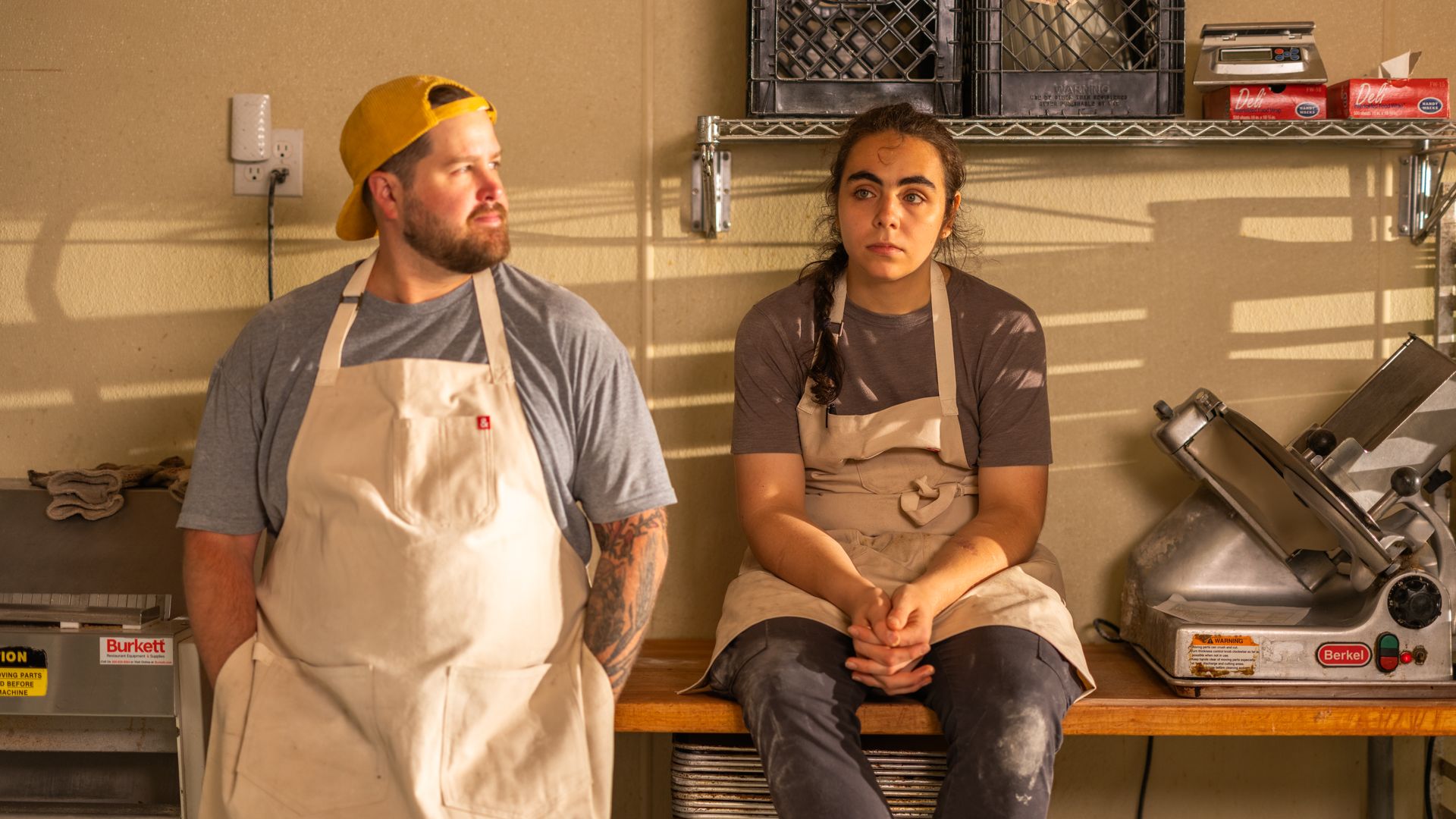 Two people in beige aprons in a kitchen workspace. One stands with a yellow cap and tattoos, looking sideways. The other sits on a wooden table, hands clasped, looking forward.