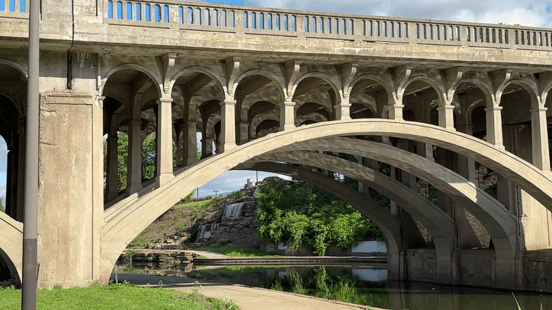 GIF shows the water reflecting light on an ornate concrete bridge and a waterfall in the background along Brush Creek.