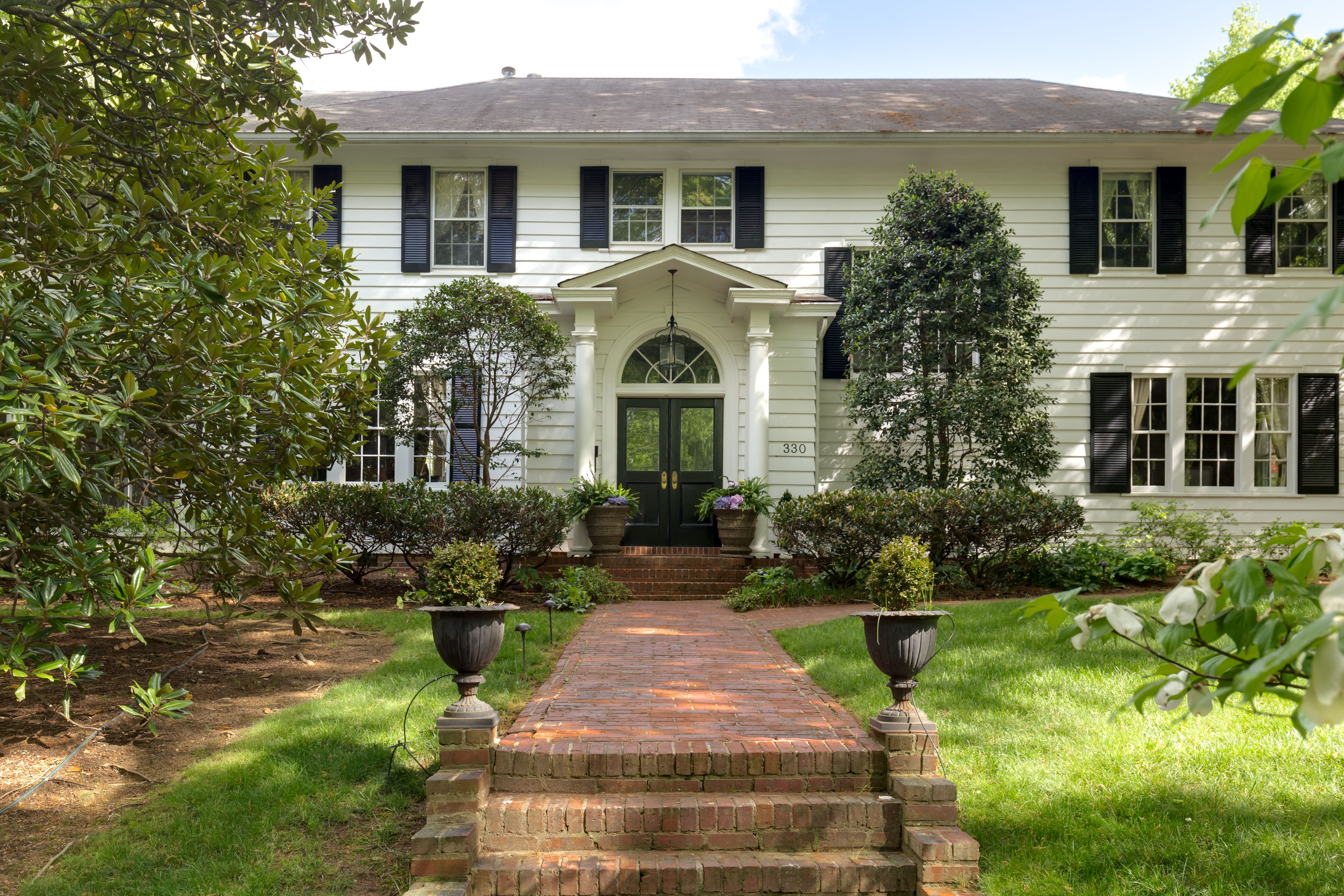 Two-story white house with black shutters, black double front doors under a small porch, brick steps and walkway, surrounded by green bushes, trees, and lawn on a sunny day.