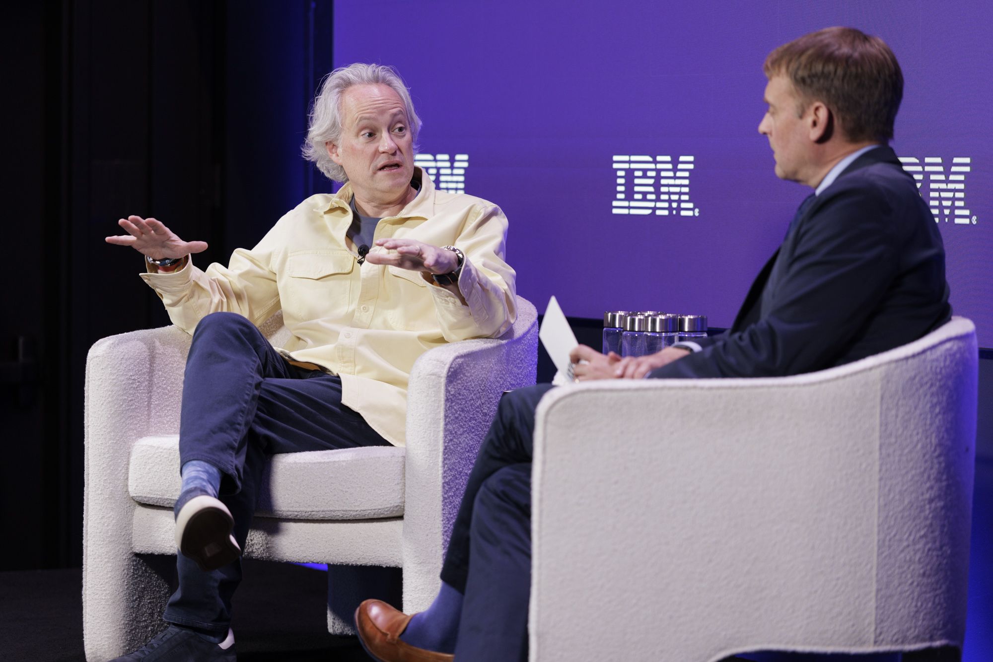 Two men sit in light armchairs on a stage with a purple IBM backdrop; the left man wears a pale yellow shirt and gestures, while the right man in a dark suit listens.