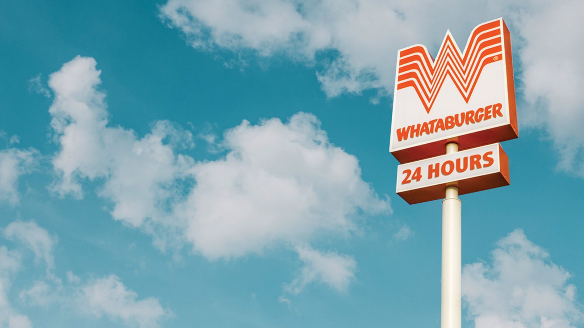 An orange-and-white sign against a blue sky with a few puffs of white clouds. The sign has a large W and, below it, "WHATABURGER 24 HOURS"