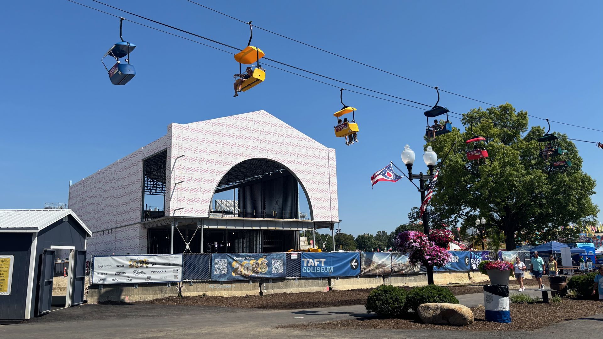 Blue sky over a fairground gondola ride: blue and yellow chairlifts suspended on cables with riders, near a white building under construction with a curved arch, with trees and people below.