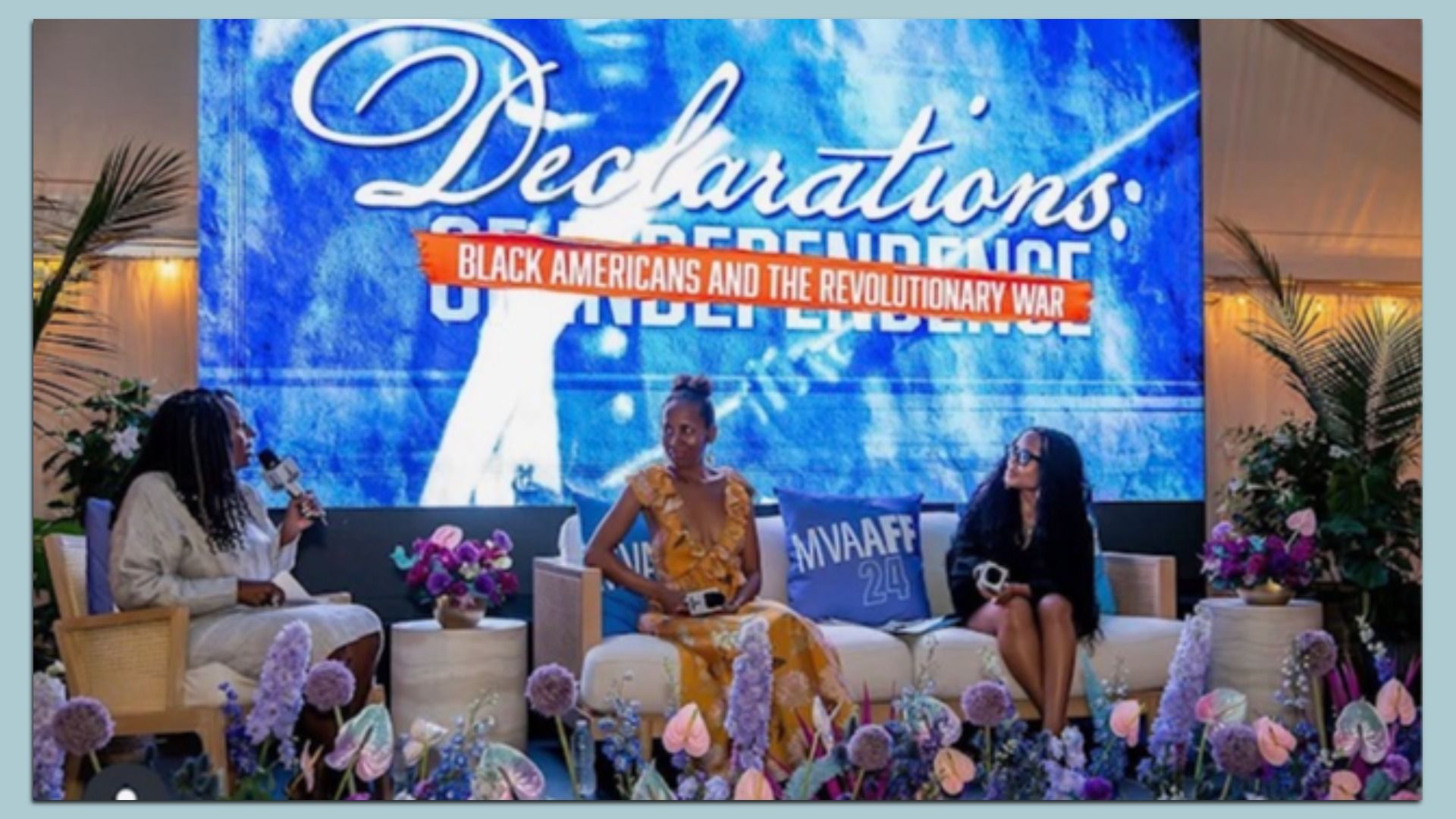 Three women sit on a panel in front of a large blue backdrop that reads "Declarations" with a red banner across it saying "Black Americans and the Revolutionary War." Purple flowers line the foreground.