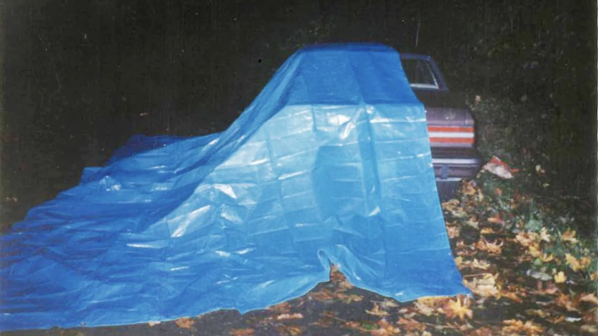 A photo of an older model car with one side draped with a blue tarp.