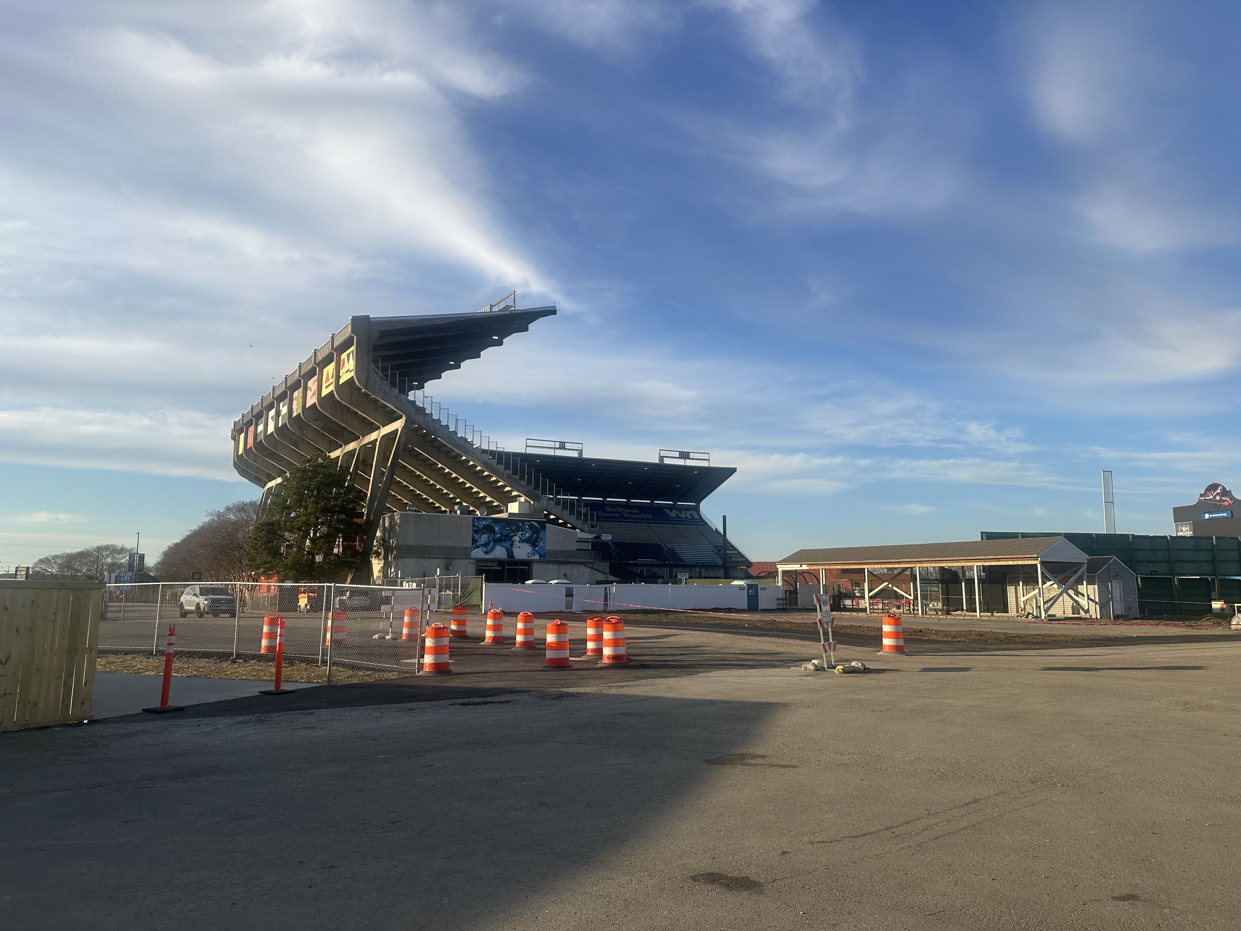 A large stadium grandstand with a cantilevered roof, banners along the rail, near a fenced construction site with orange traffic cones; a clear blue sky above.