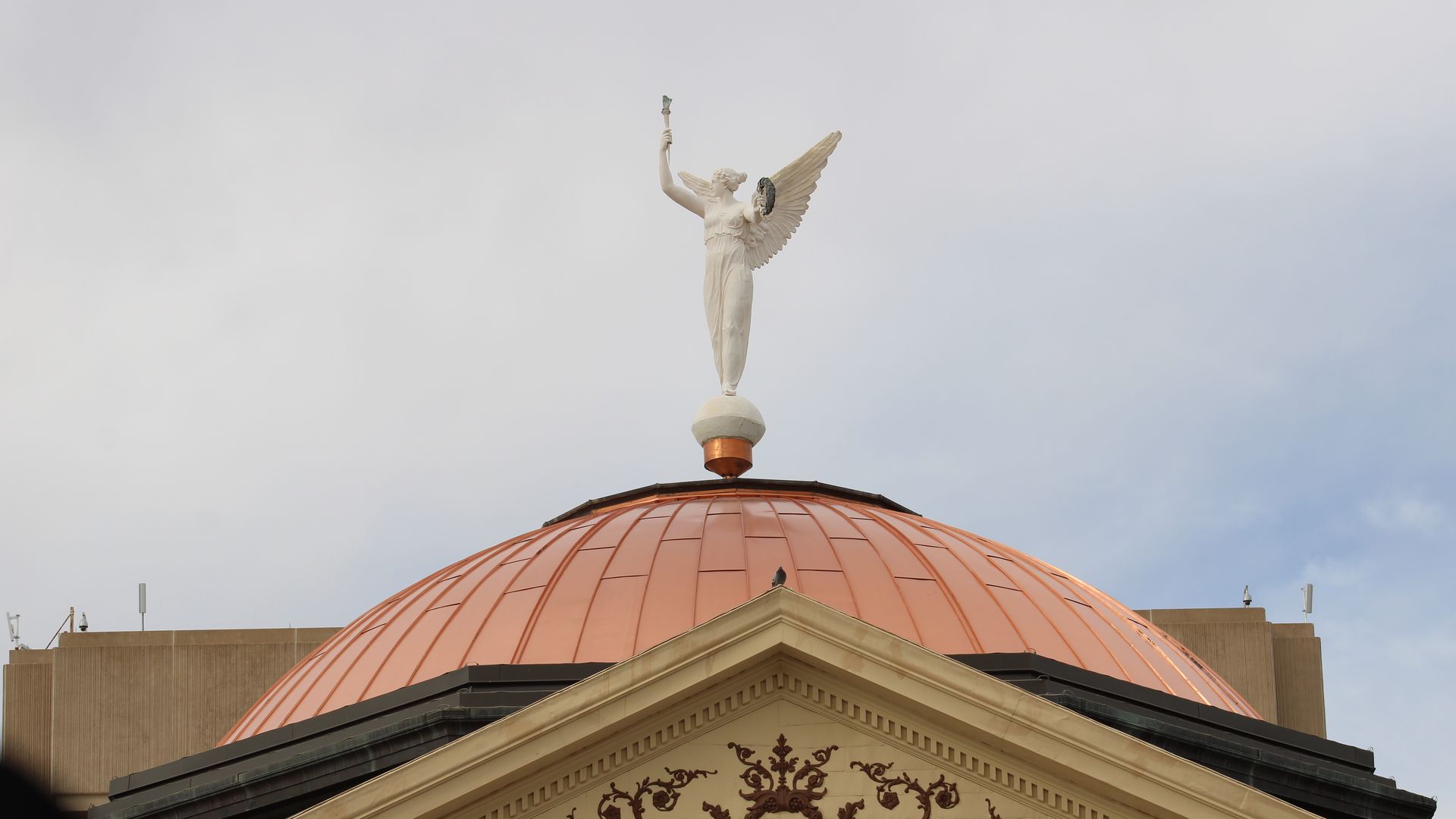A white statue of a winged woman holding a torch atop a copper dome. 