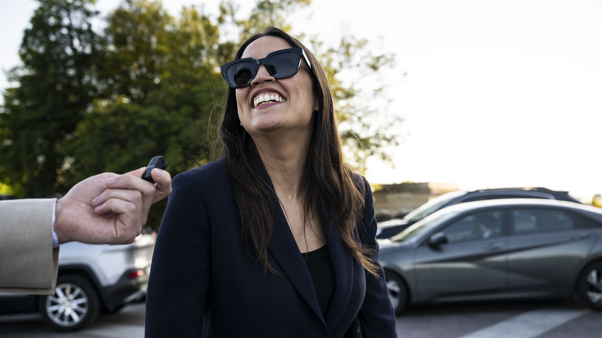 Rep. Alexandria Ocasio-Cortez, wearing sunglasses, smiles as she's asked a question by a reporter with a microphone.