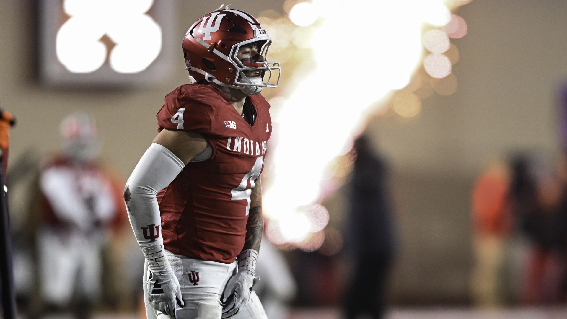 Football player wearing a red Indiana jersey and helmet with number 4, standing on the field with bright fireworks in the background during a night game.