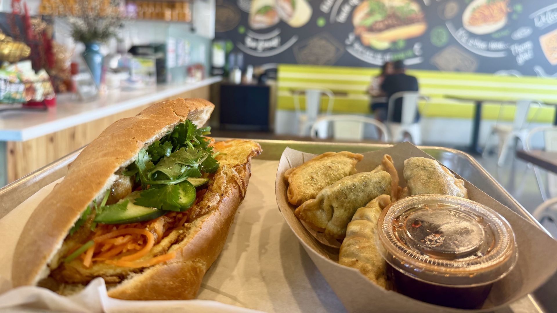 Close-up of a Vietnamese banh mi sandwich with cilantro and pickled veggies on a crusty baguette, on a counter; blurred chalkboard mural and pastries in the background.