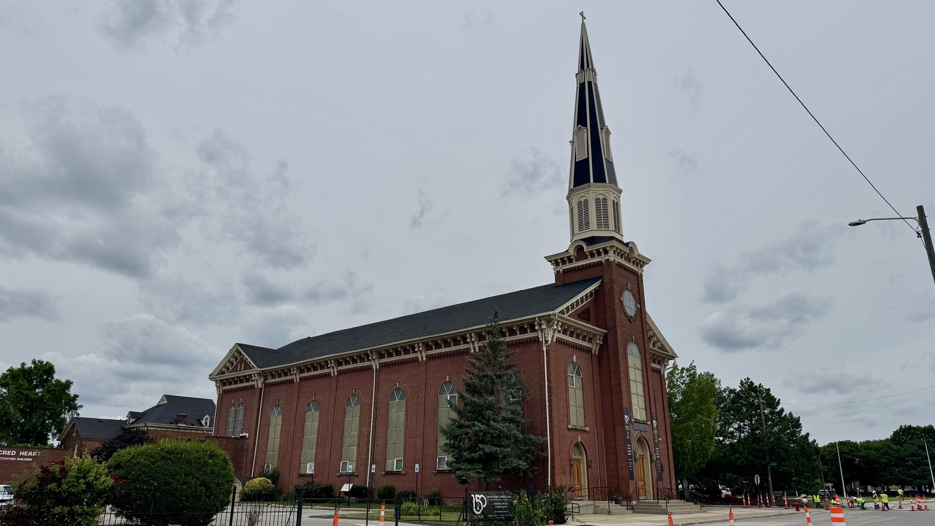 A red brick church with a tall steeple and white trim rises above arched windows under a cloudy sky.