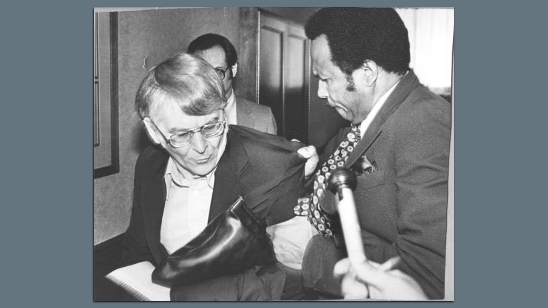 Black and white photo of a black man muscling a bespectacled white man out of a room.