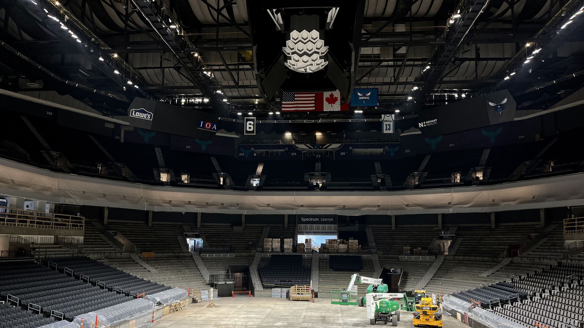 Empty indoor arena under construction or renovation, with covered seats, construction equipment on concrete floor, and flags including US and Canada hanging above.