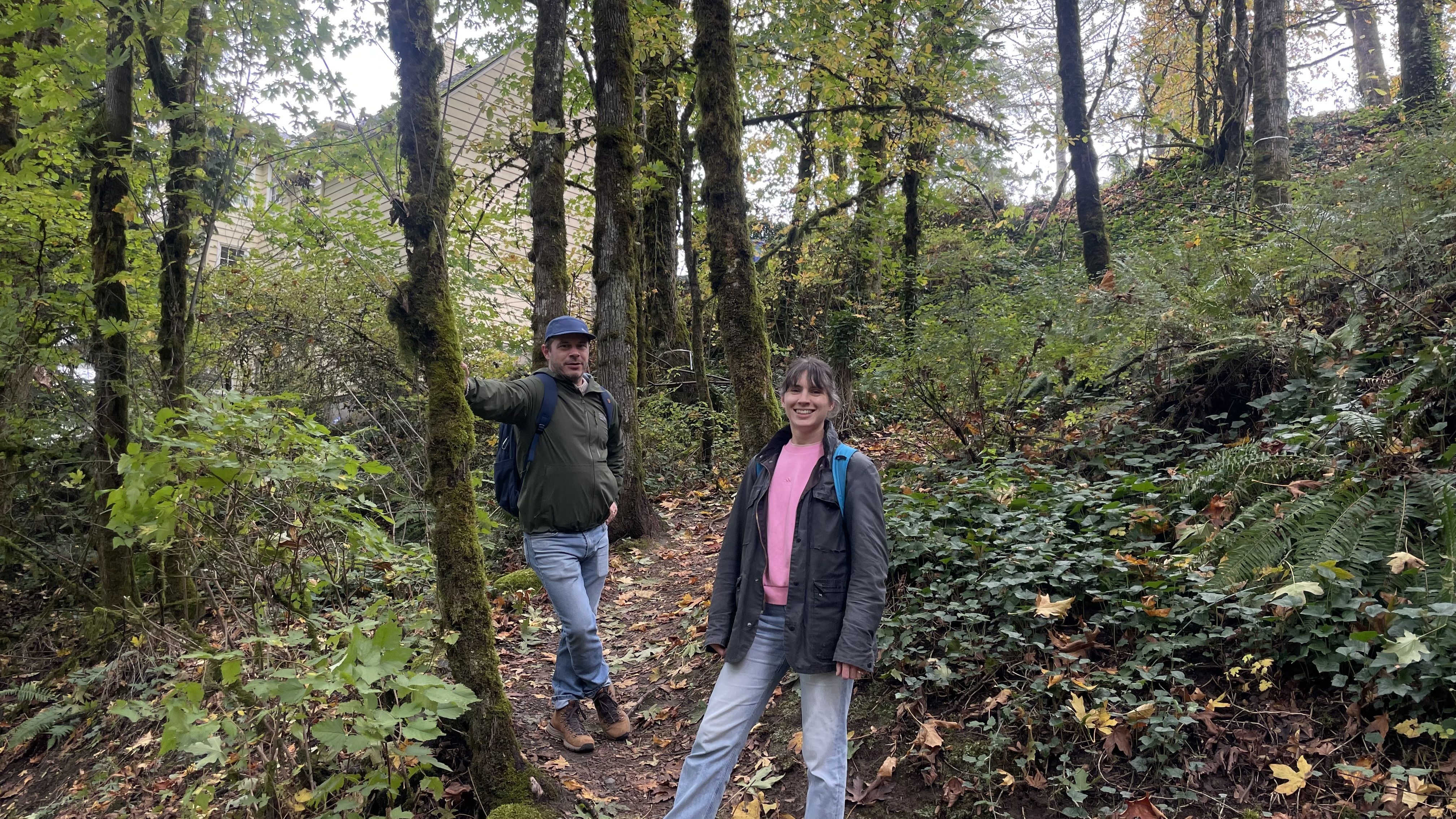 Two people wearing jackets and backpacks stand on a forest trail surrounded by green foliage and trees with a beige house partially visible in the background.