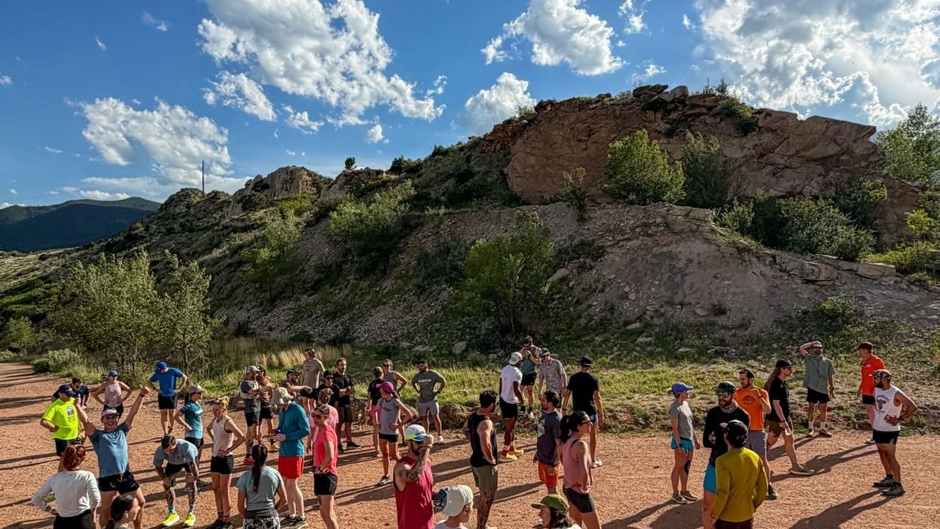Group of people in bright athletic gear standing on a reddish dirt trail at the base of rocky hills, under a blue sky with white, fluffy clouds.