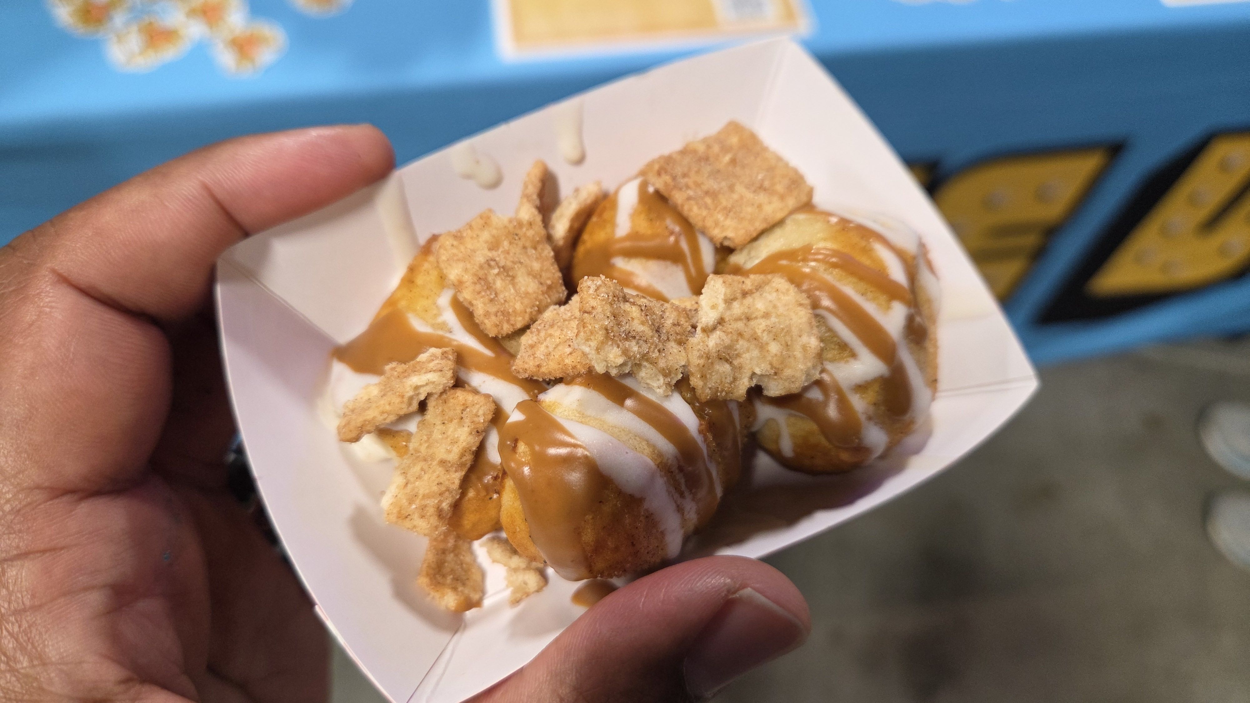 Hand holding a small paper tray with three glazed doughnut holes topped with caramel and white icing, garnished with cinnamon cereal pieces.