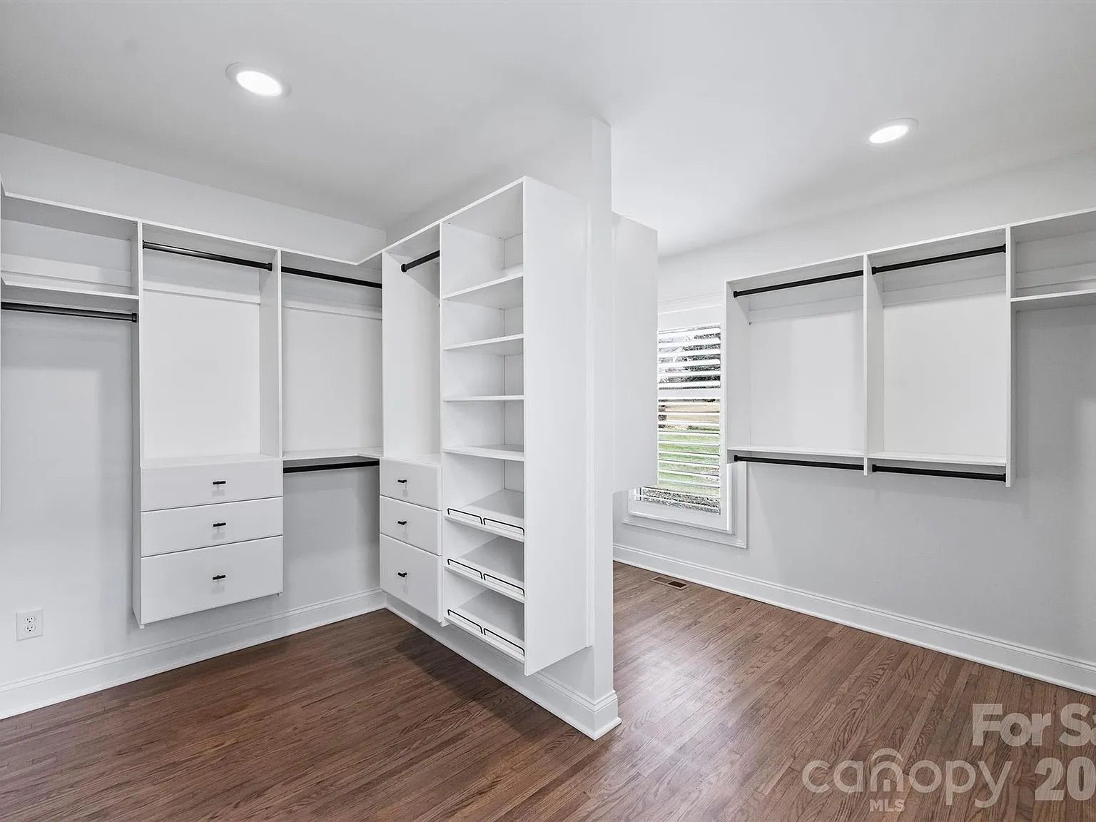 Spacious walk-in closet with white built-in shelves, drawers, and hanging rods. Wooden floor and a window with white blinds letting in natural light.