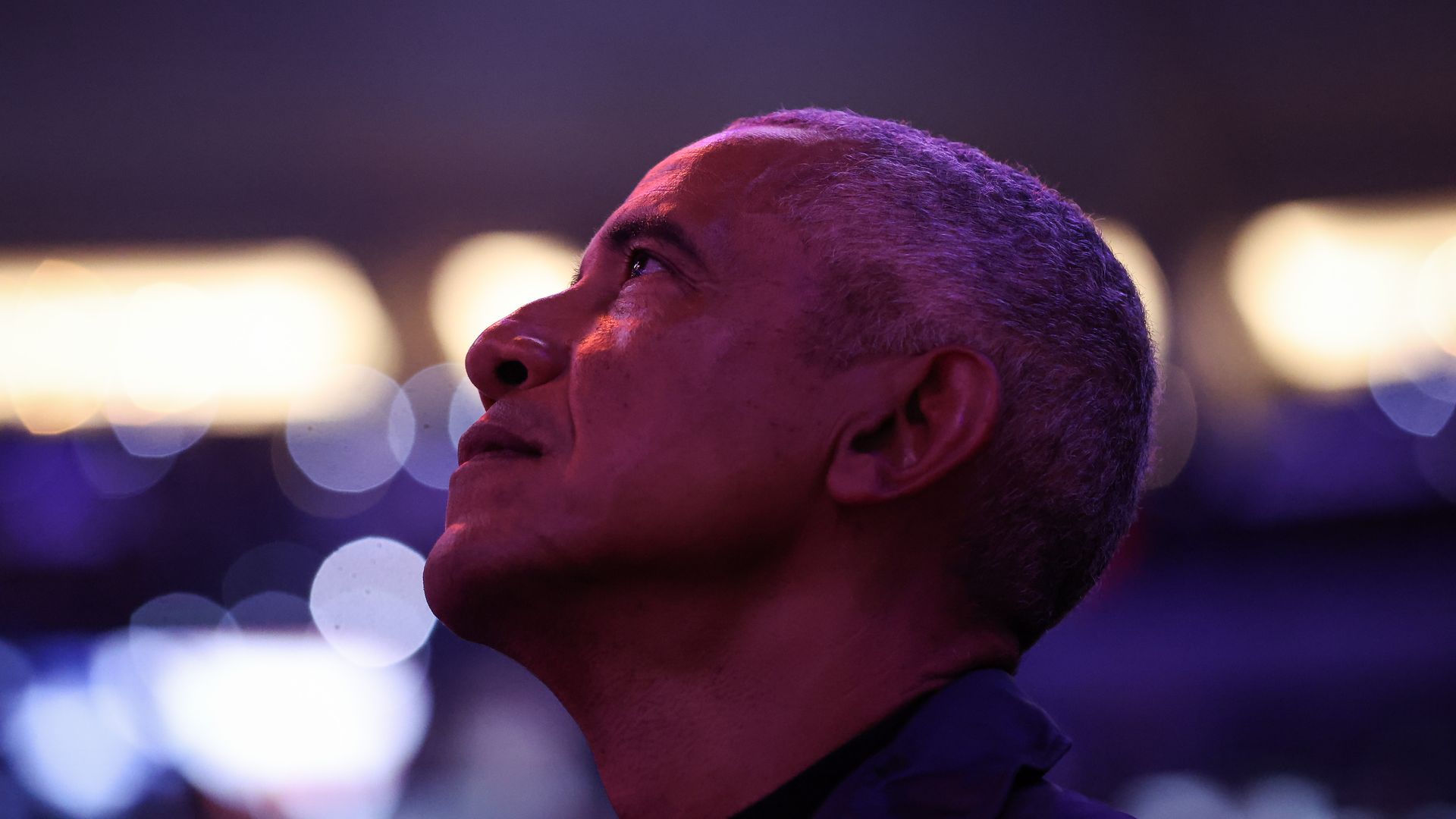 Barack Obama, in a photo taken from the side with a blurred background, looks up in a stadium during a basketball game.
