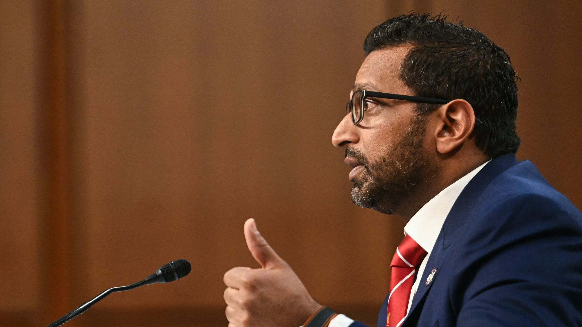 Kash Patel holds his thumb up as he speaks into a microphone wearing glasses, a red tie and a blue suit.