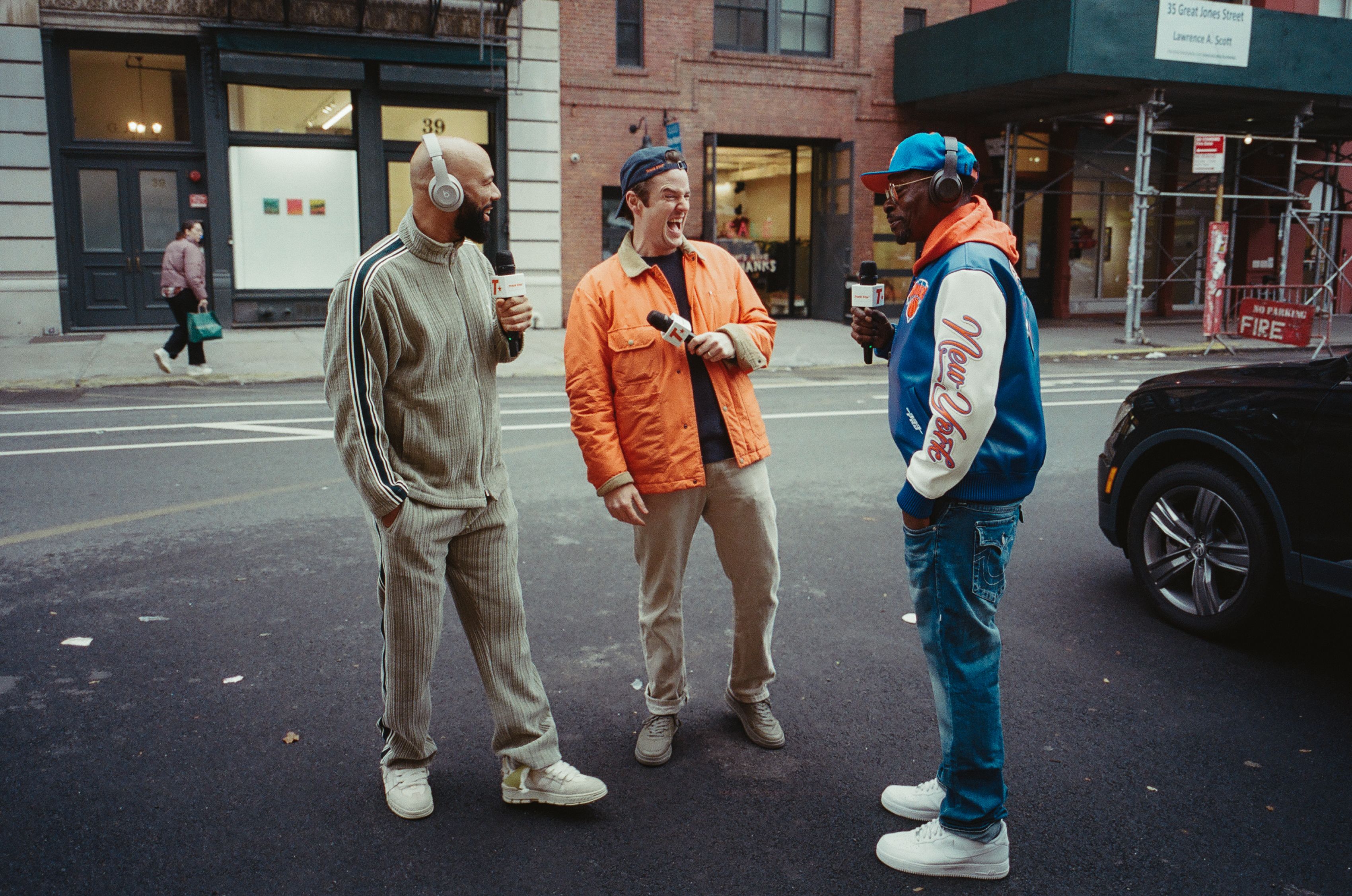 Three men standing on a city street, holding microphones and wearing headphones, dressed casually in gray tracksuit, orange jacket, and New York Knicks jacket and blue cap, chatting and laughing.