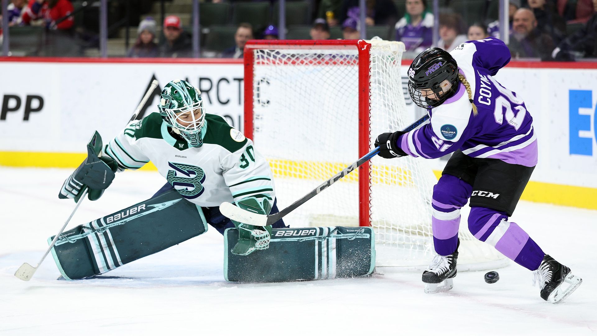A hockey player in a purple jersey swings her stick at a puck as a goaltender in pads defends the net during a game